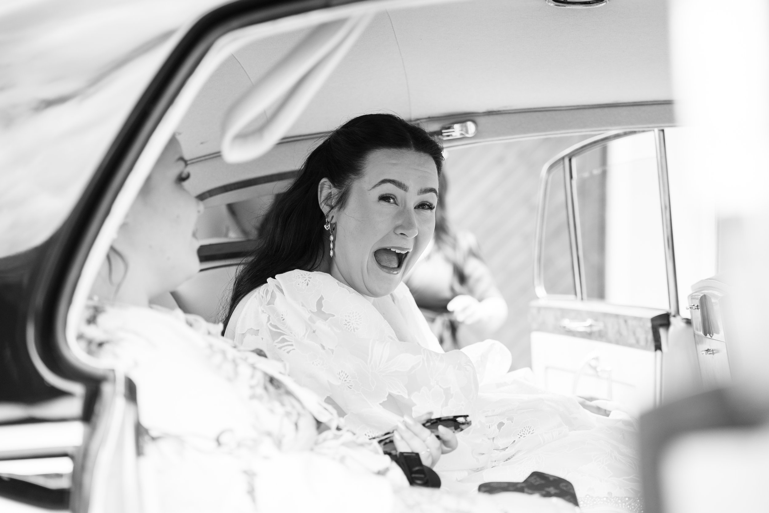 A woman with dark hair and earrings, wearing a white dress, sitting inside a vintage car, making a surprised or excited facial expression.