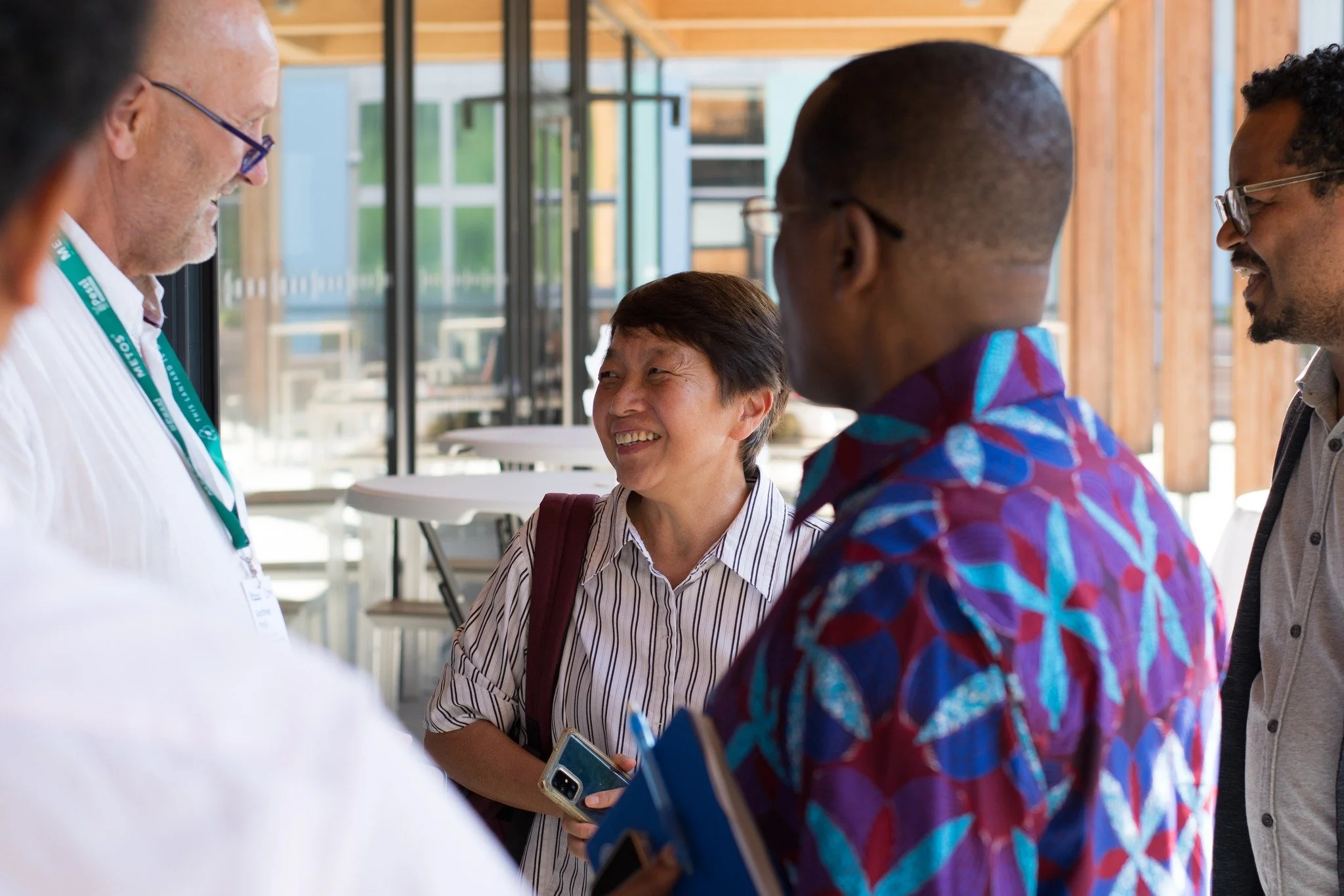 Group of five people, diverse in ethnicity, smiling and talking indoors with large windows and wooden walls.