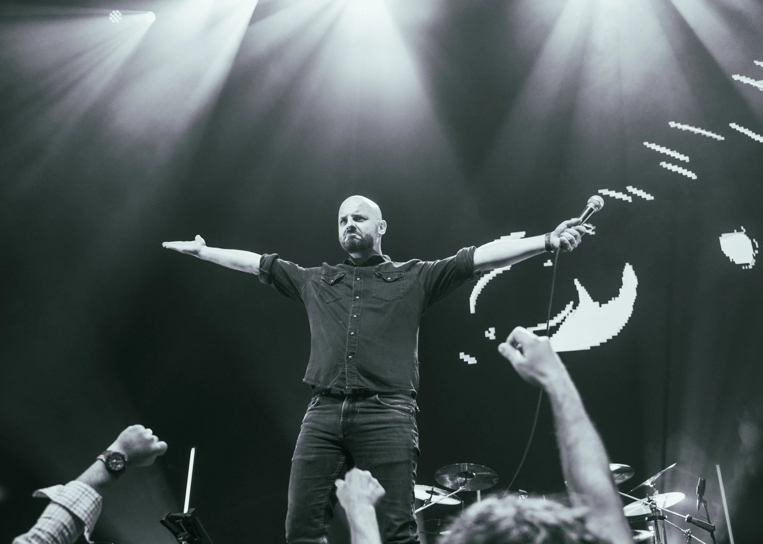 A performer on stage with arms outstretched, holding a microphone in one hand, facing an audience with fists raised. The stage is lit with bright spotlights and a large digital screen with pixelated graphics is in the background.