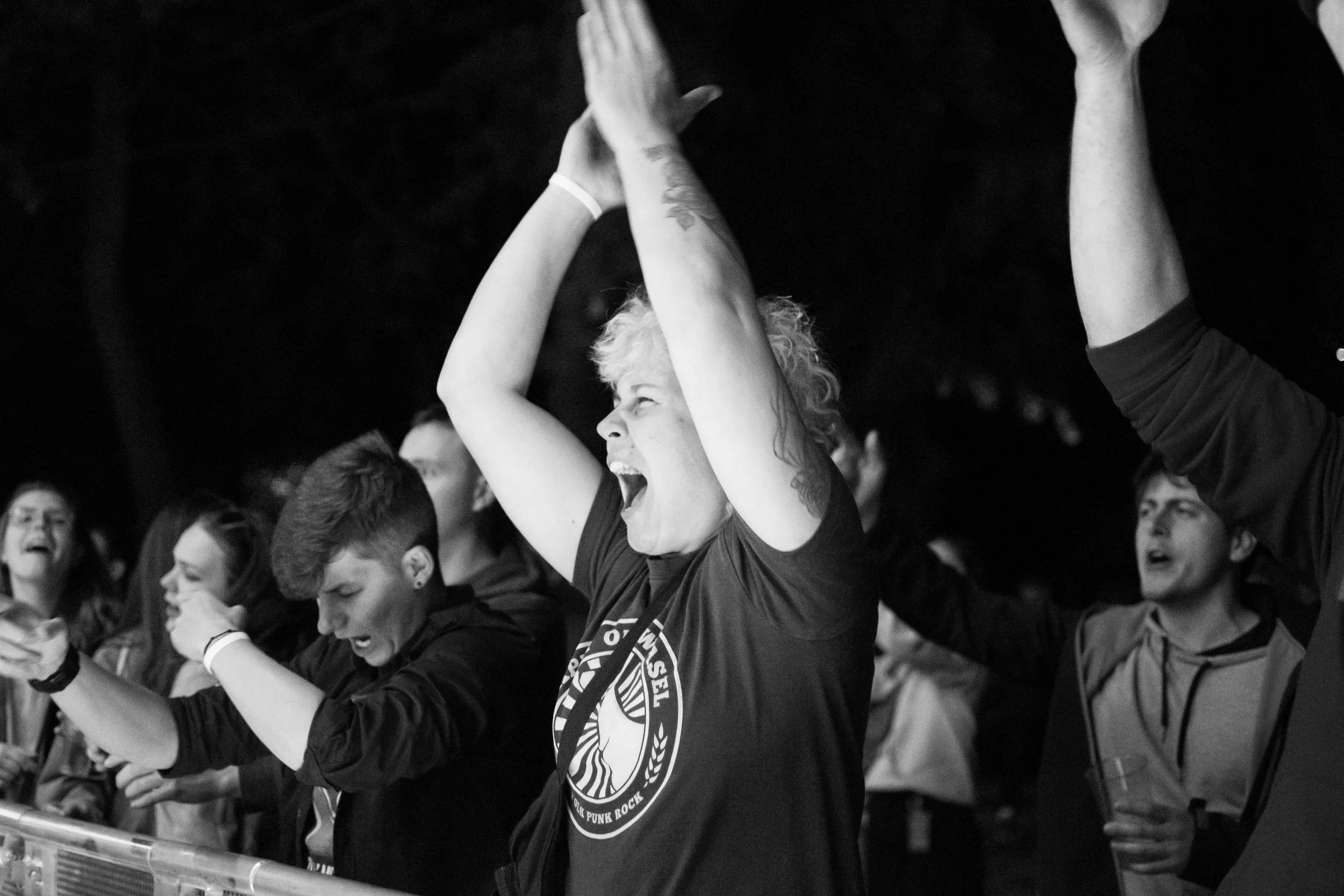 Group of people at concert or event, some with arms raised, celebrating or cheering, in black and white.