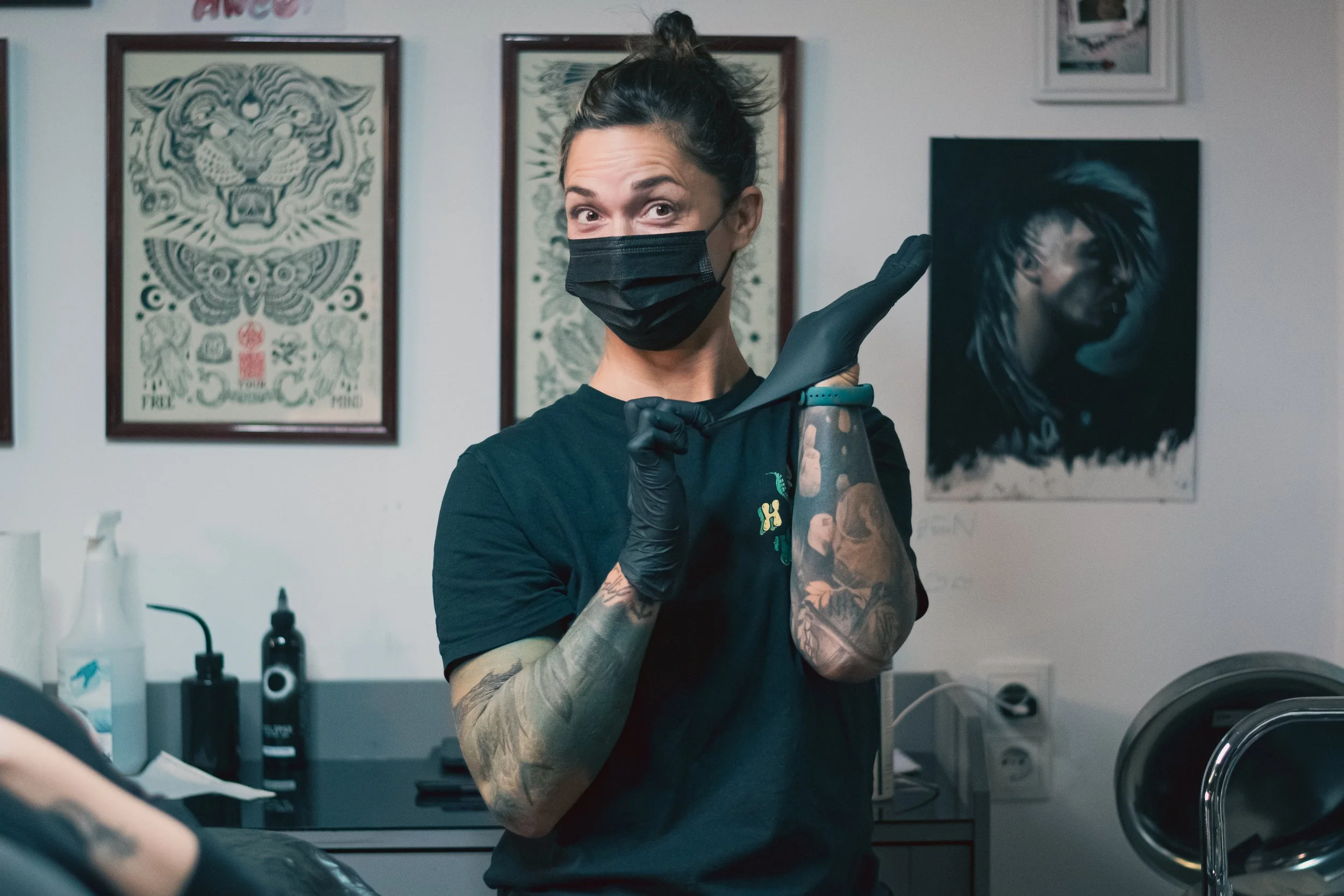 A tattooed woman wearing a black face mask and black gloves is preparing to tattoo a person's arm in a tattoo studio.