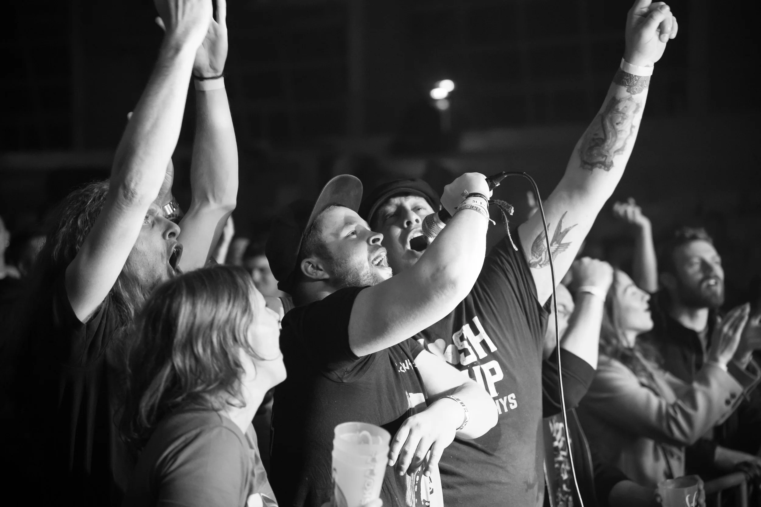 People at a concert or rally, some singing or shouting, raising their hands, with one man holding a microphone and wearing a t-shirt that says 'Close the Gap,' standing among other enthusiastic crowd members.