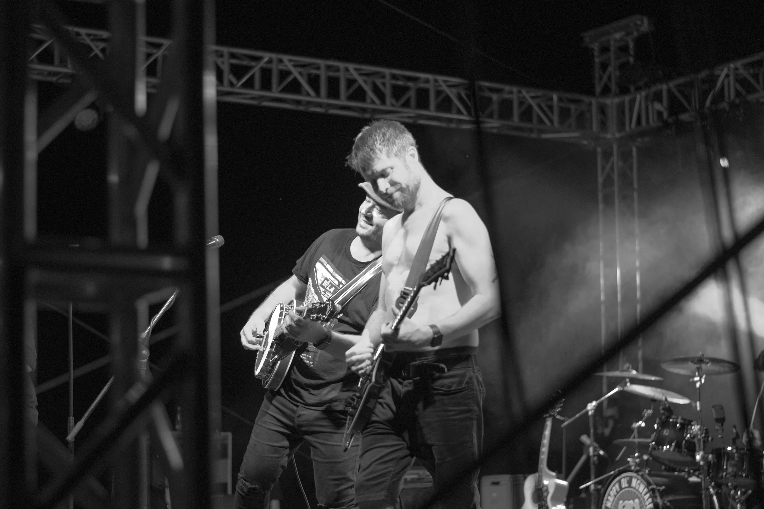 Two male musicians playing guitars on stage during a concert, with stage equipment and drums in the background, black and white photo.