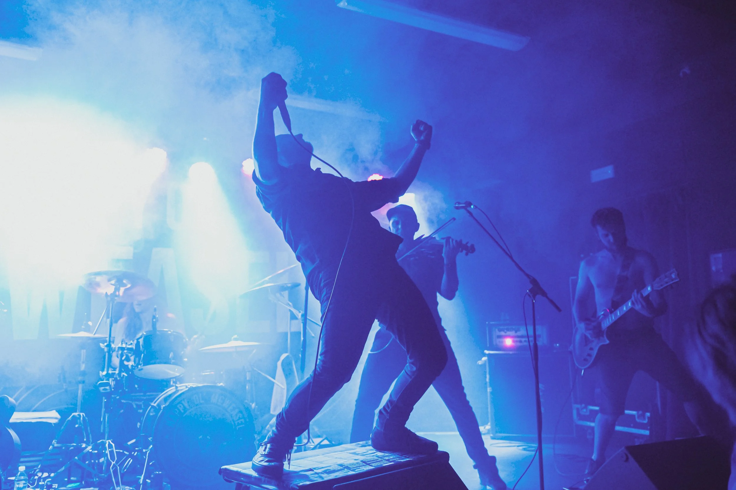 Band performing on stage with blue lighting, one musician standing on a platform with a microphone, another playing violin, and a third playing electric guitar.