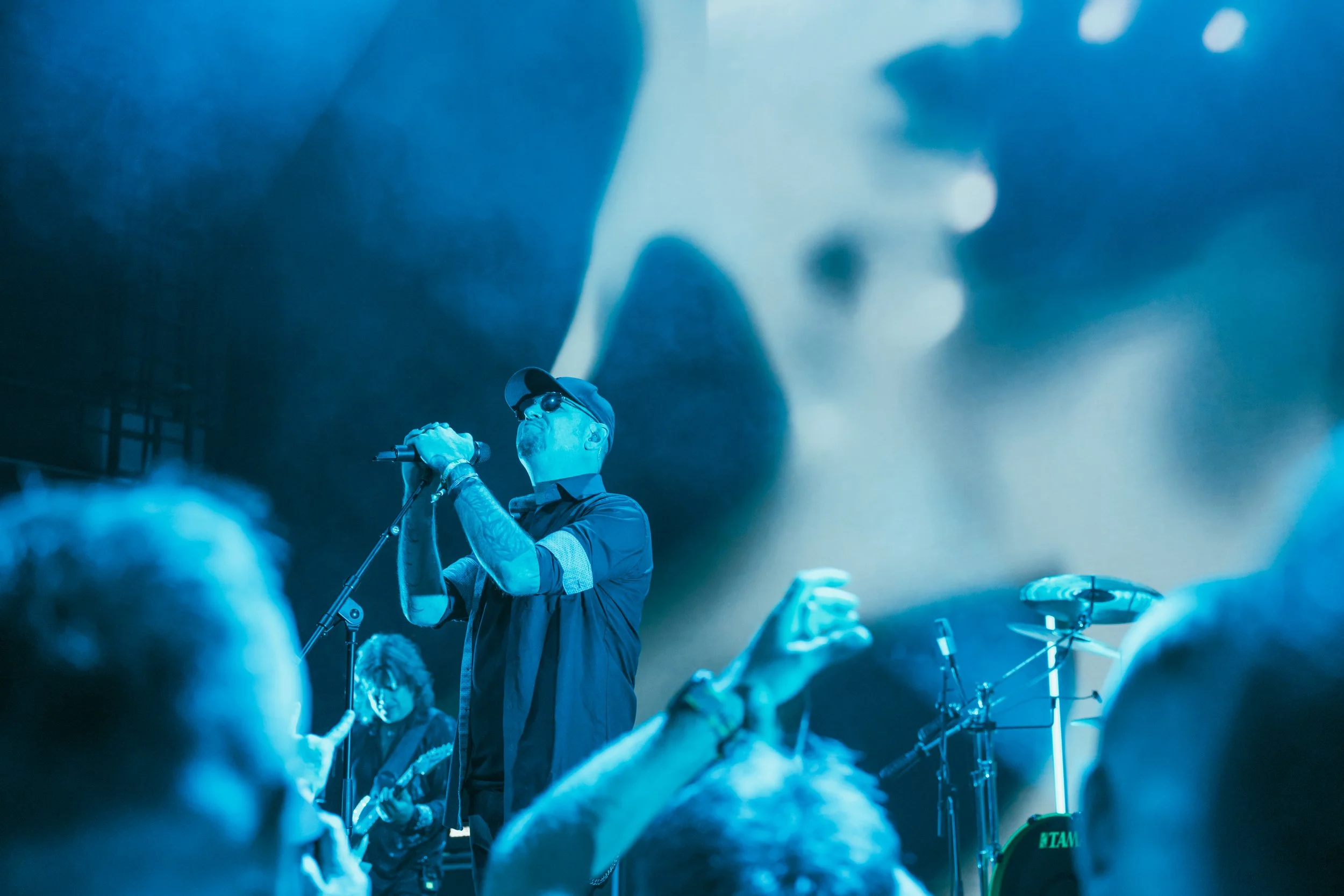 A male singer wearing sunglasses, a cap, and tattoos, singing into a microphone on stage surrounded by an audience, with a musician playing guitar and a large screen in the background displaying visuals.