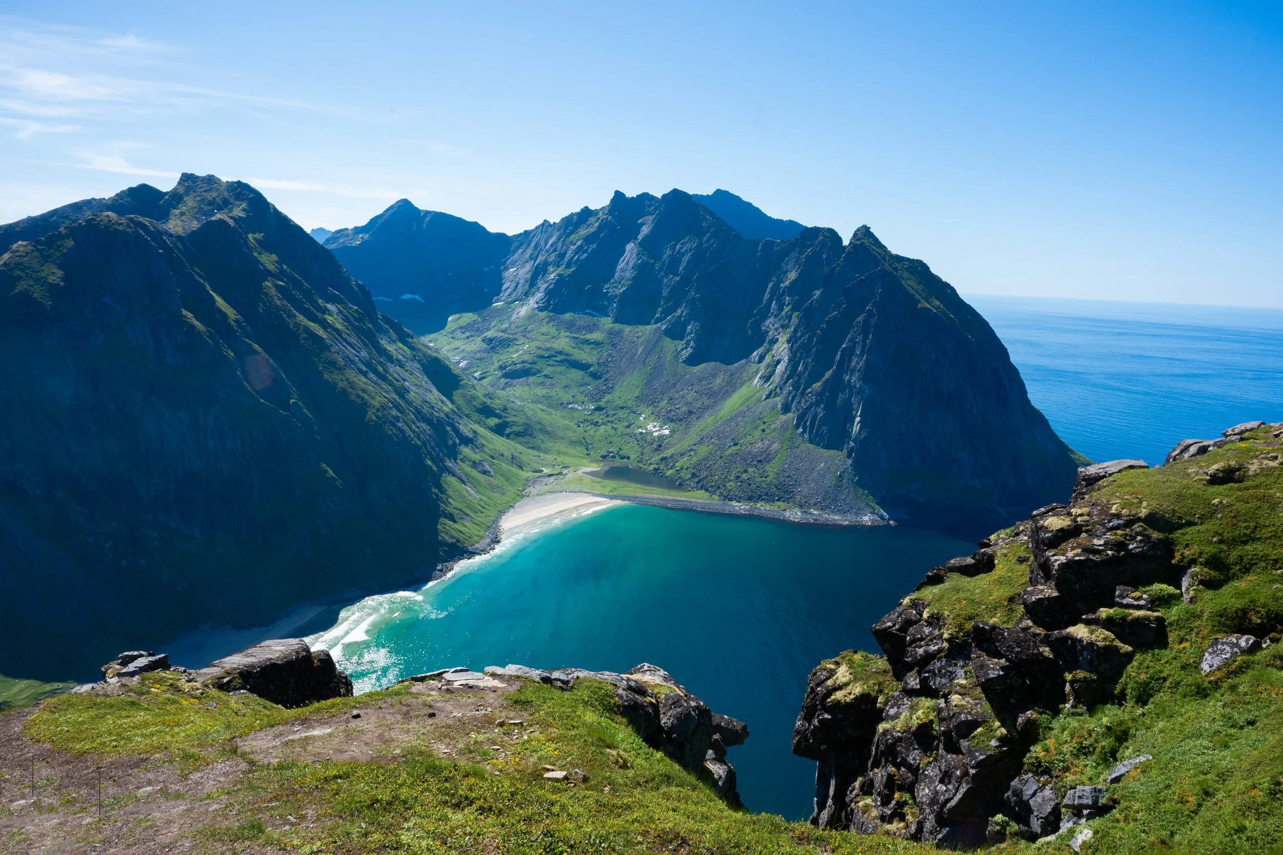 A panoramic view of a green mountainous landscape with a steep-sided valley that contains a small beach and a turquoise lake, with the ocean visible in the distance under a clear blue sky.
