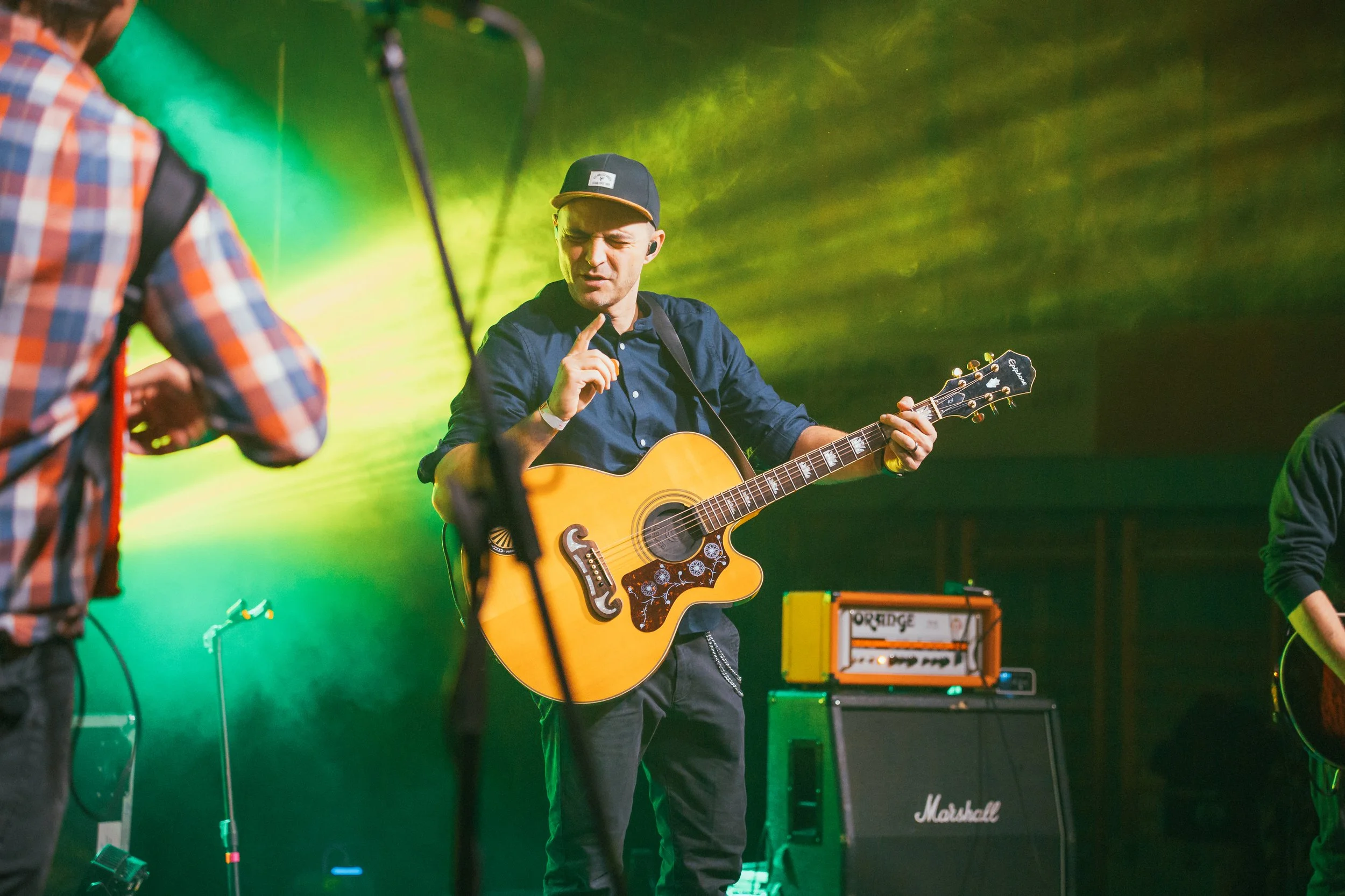 A man playing an acoustic guitar on stage with green and yellow lighting, wearing a black cap, dark shirt, and black pants, making a hand gesture, with musical equipment in the background.