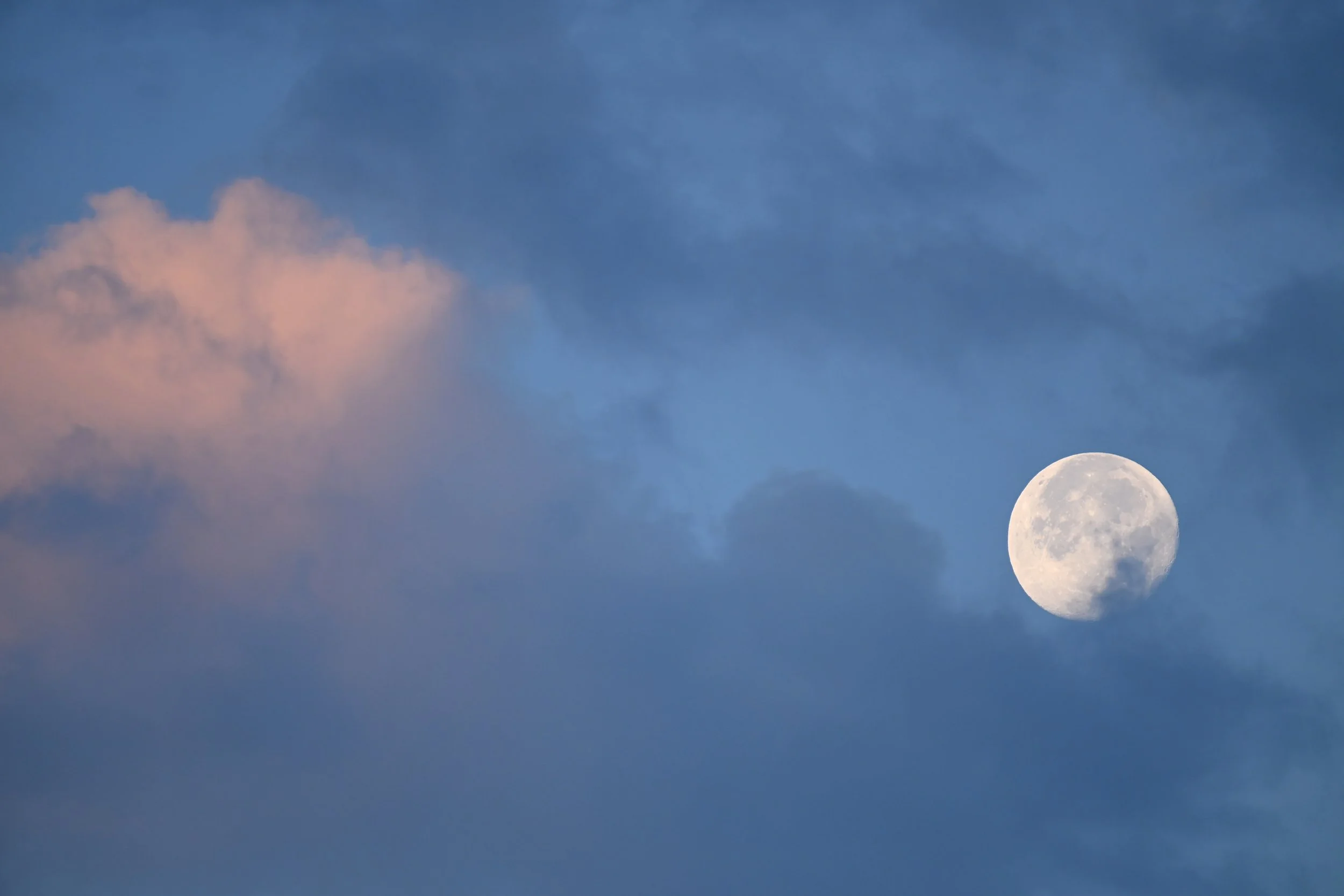 The image shows the moon in a partly cloudy sky during evening or early night, with pink-tinged clouds on the upper left.