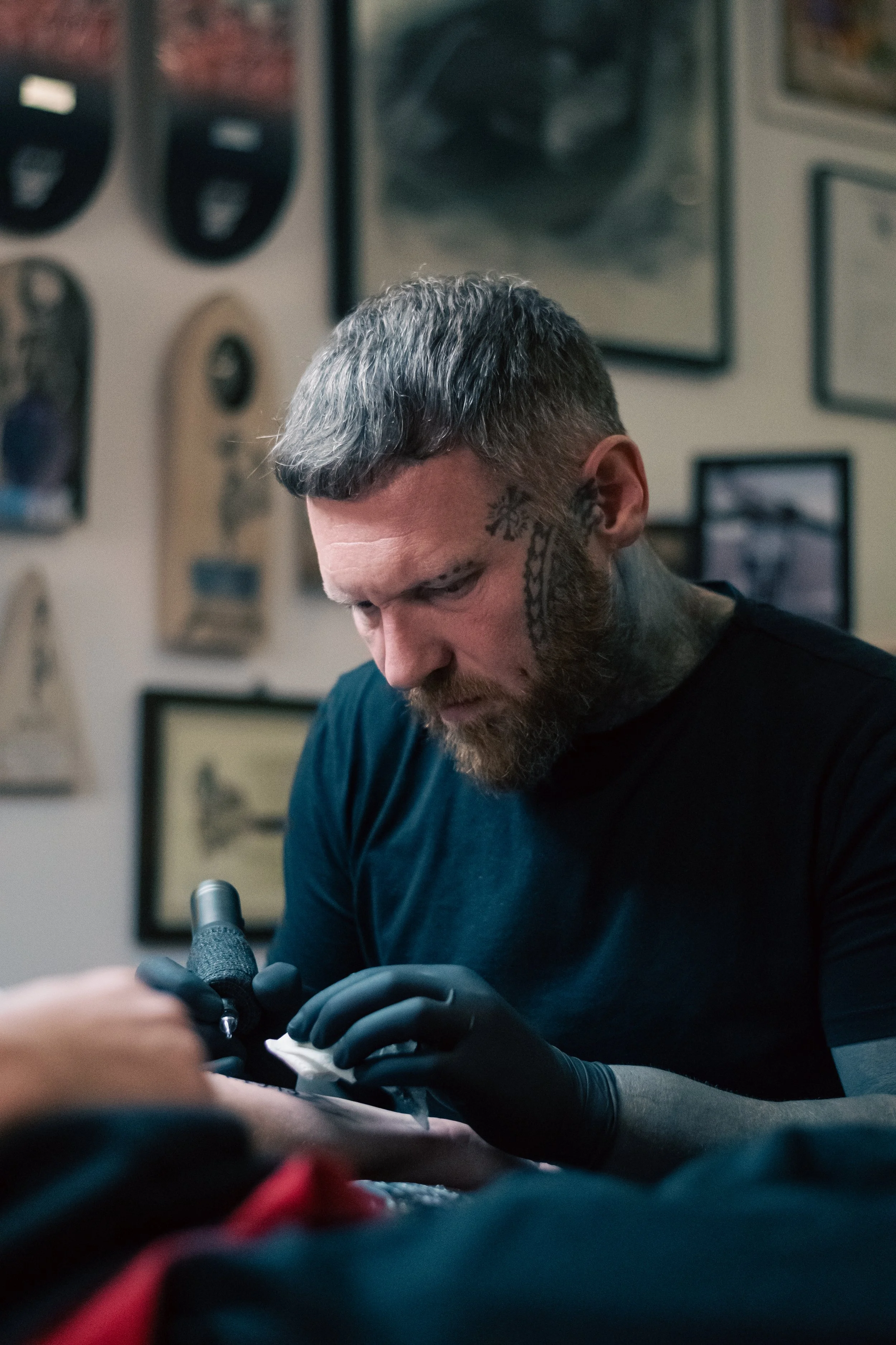 A man with tattoos and a beard, wearing black gloves, is working on a tattoo machine in a tattoo studio surrounded by framed pictures and artwork on the wall.