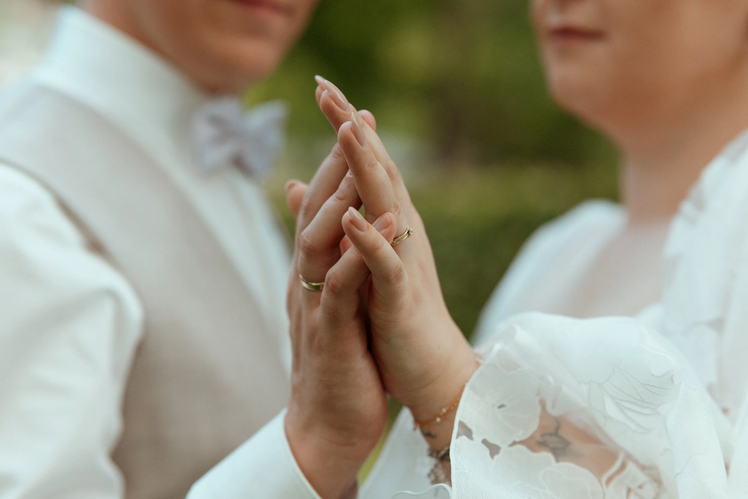 Close-up of two people holding hands, wearing wedding rings, with their palms pressed together in an intimate gesture, dressed in wedding attire.
