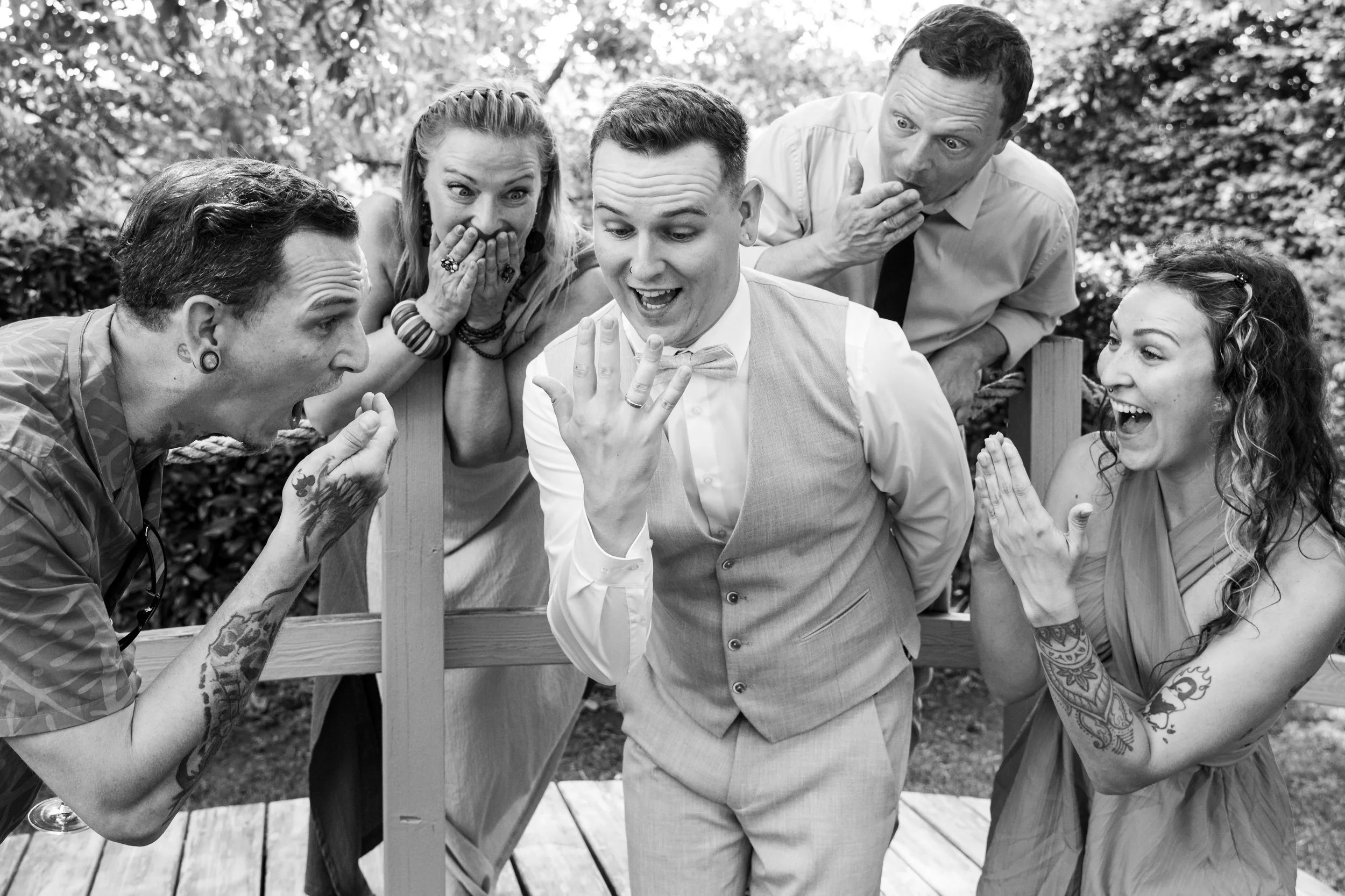 Group of six friends outdoors celebrating a wedding, with the groom showing off his wedding ring, while friends display excitement and surprise.