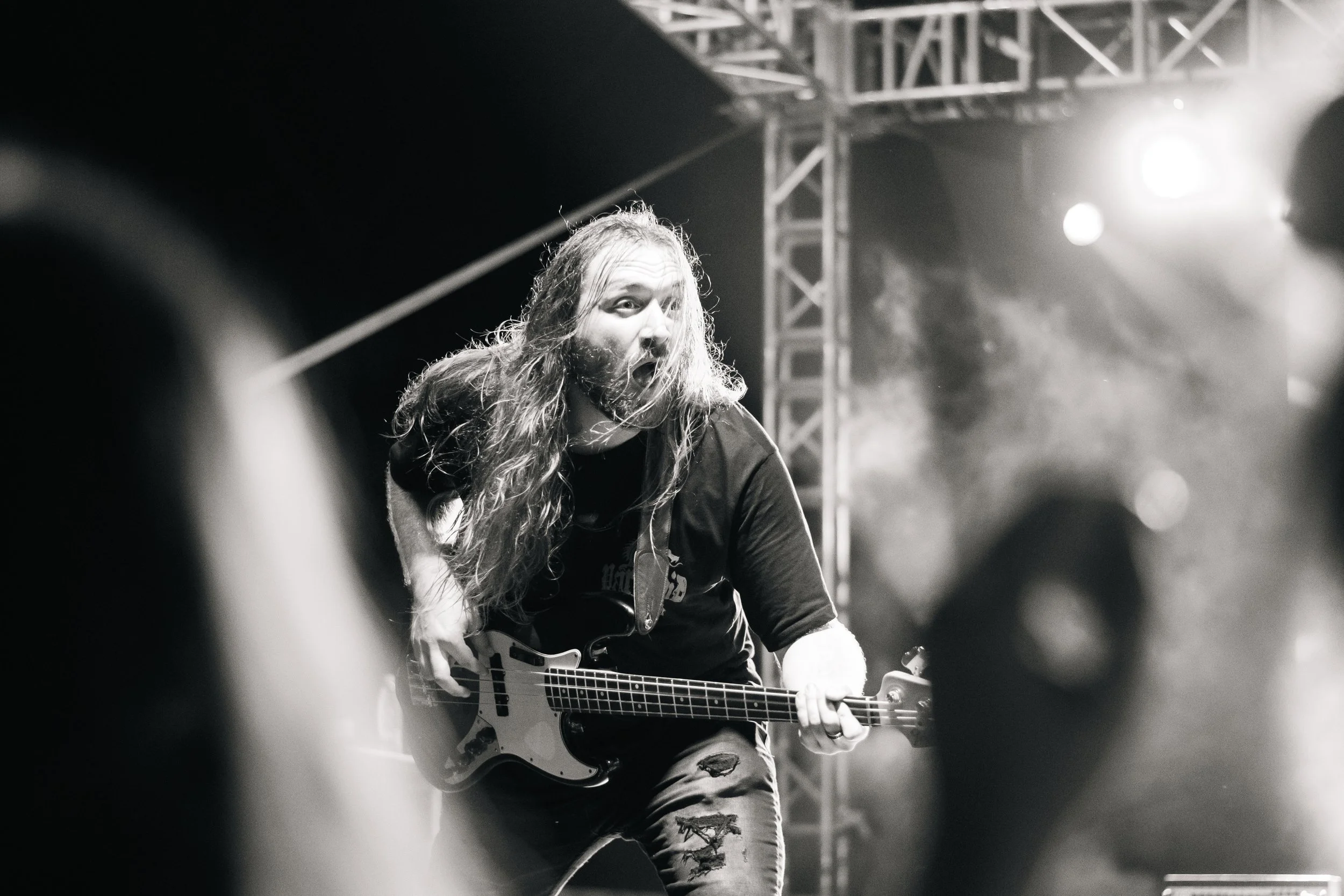 A male guitarist with long hair and beard playing an electric guitar on stage during a concert, with stage lights and structural trusses in the background.