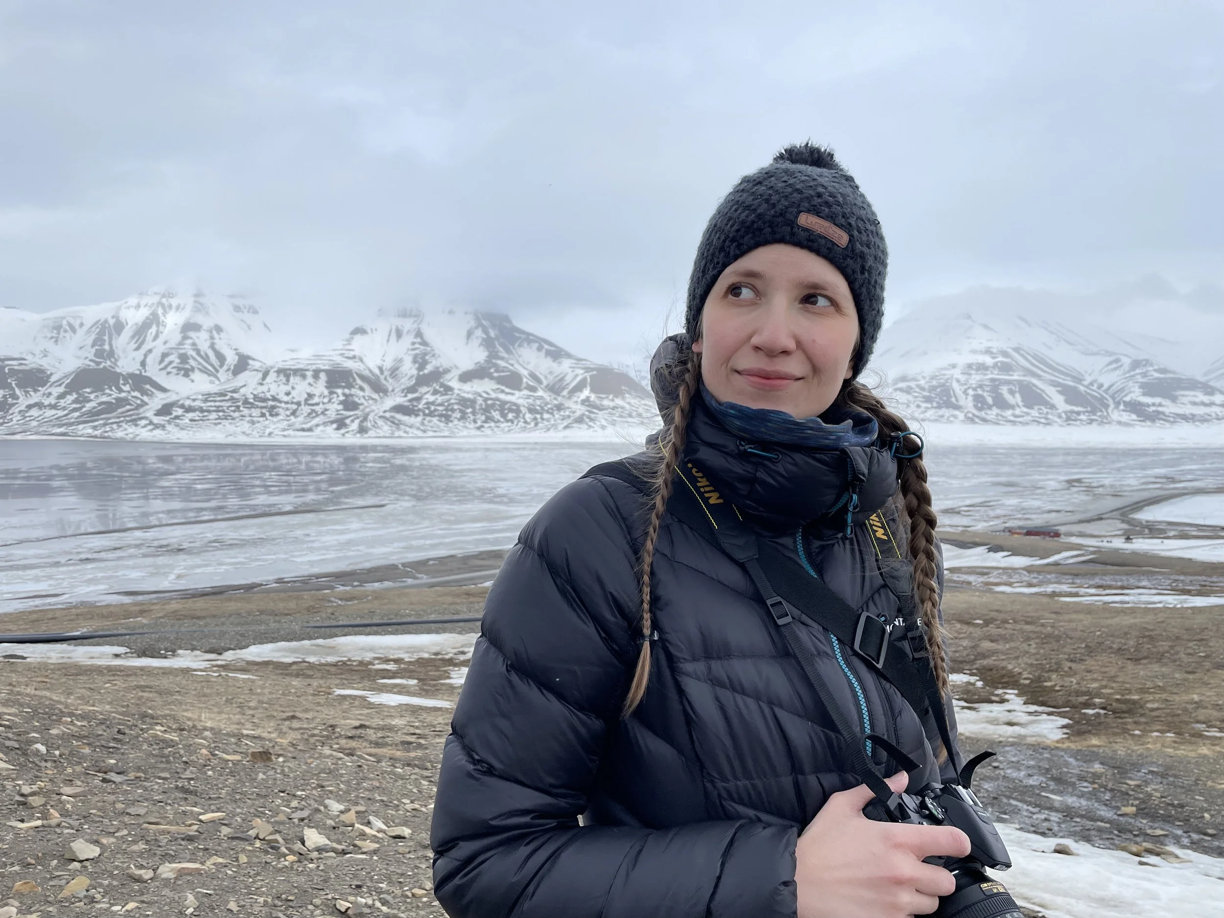 A woman in a black down jacket and knit beanie holding a camera, standing on a rocky shore with snowy mountains and icy waters in the background.