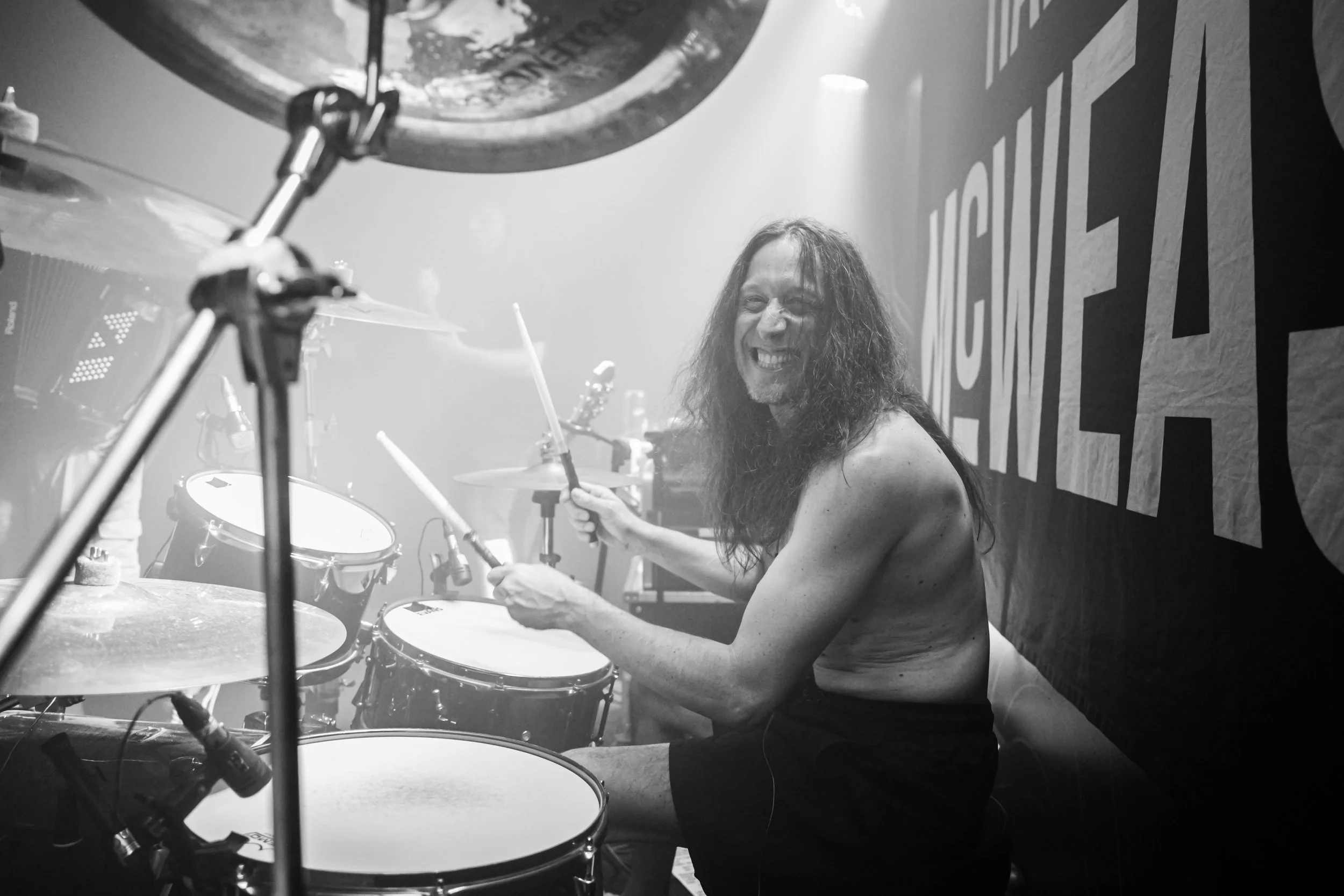 A man with long curly hair smiling while playing drums in a music venue or rehearsal space, with a large banner or sign behind him.