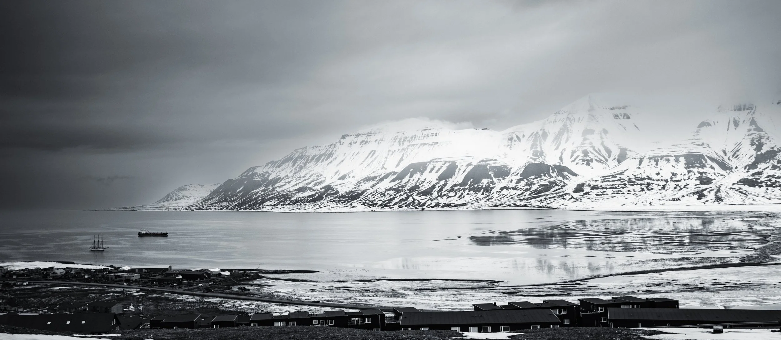 Snow-capped mountains overlooking a calm body of water with a small village and ships in the foreground, under cloudy skies.