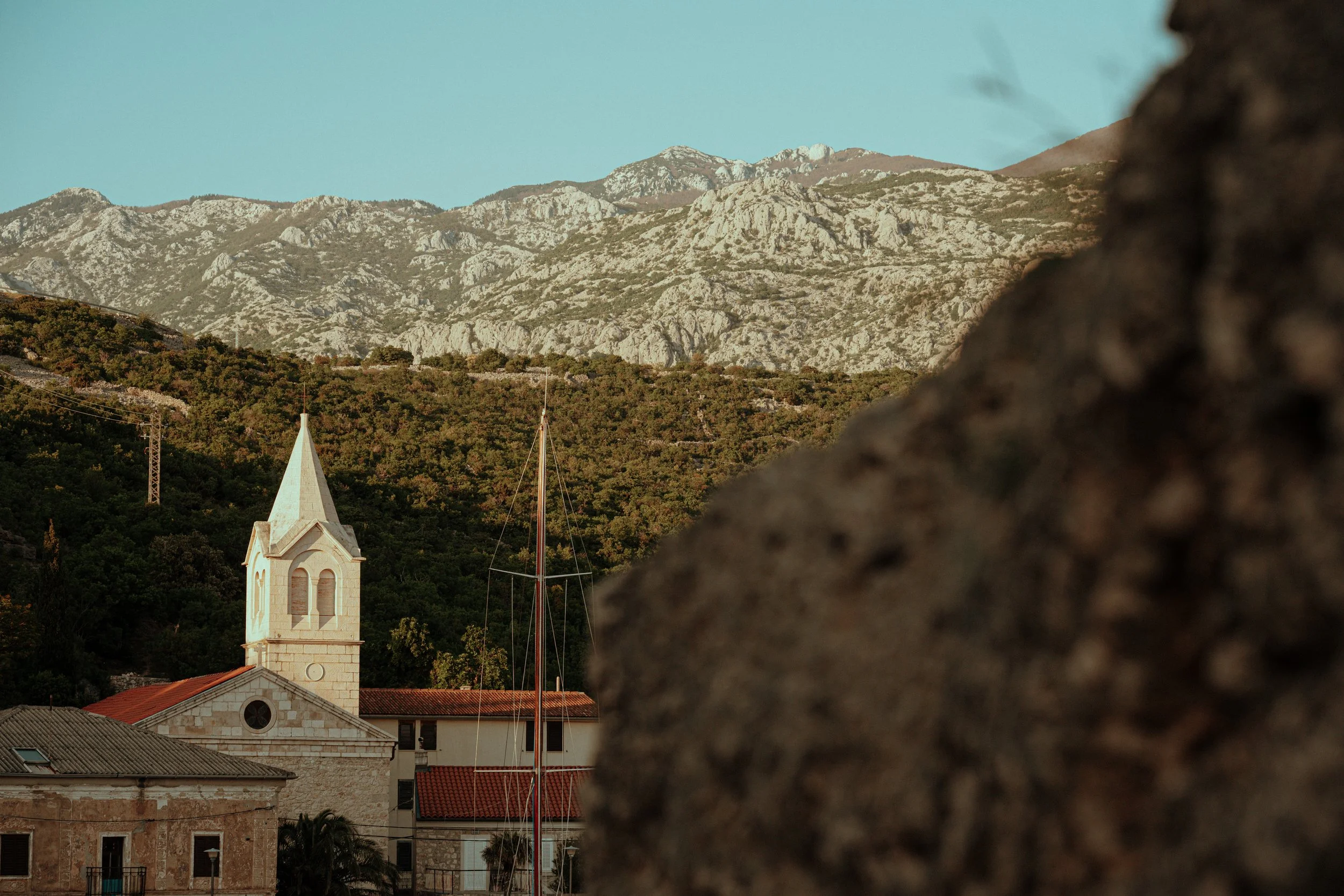 A town with a church featuring a tall spire, surrounded by a mountain range with rocky peaks, on a clear day.