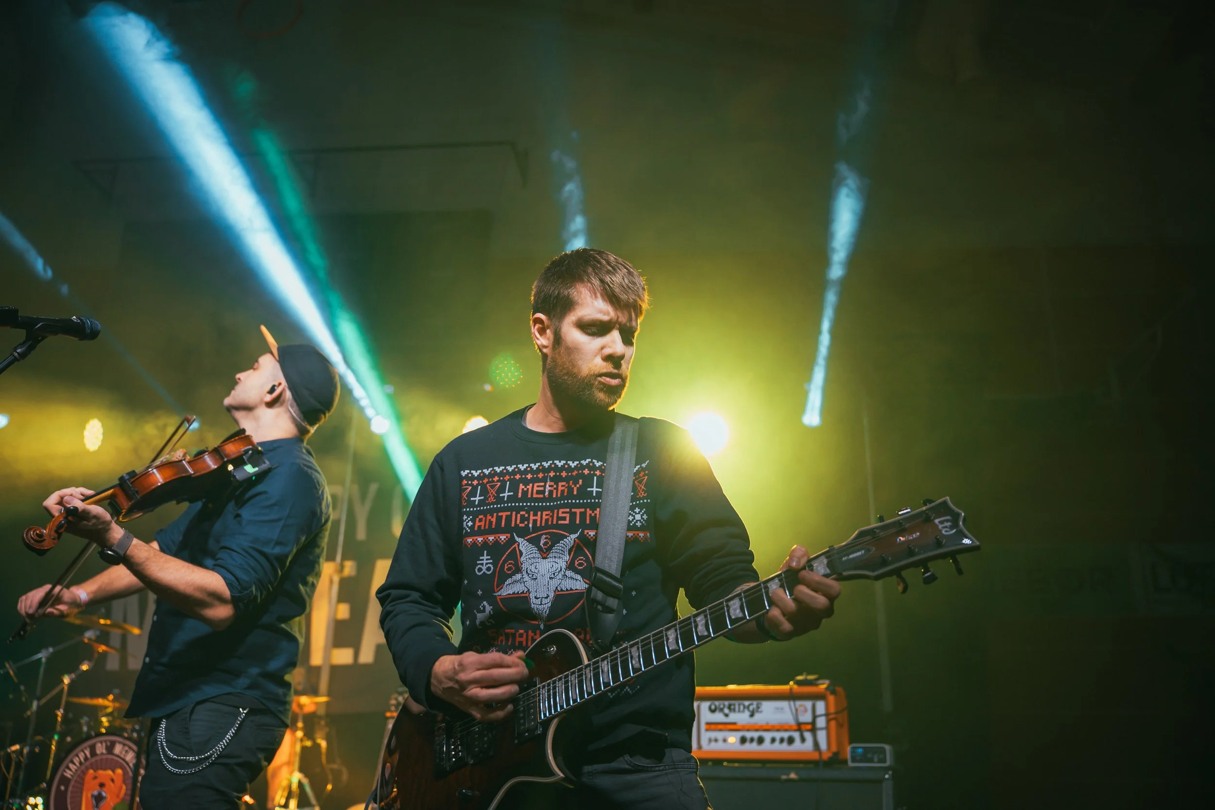 Two musicians performing on stage with colorful lighting, one playing electric guitar and the other playing violin.