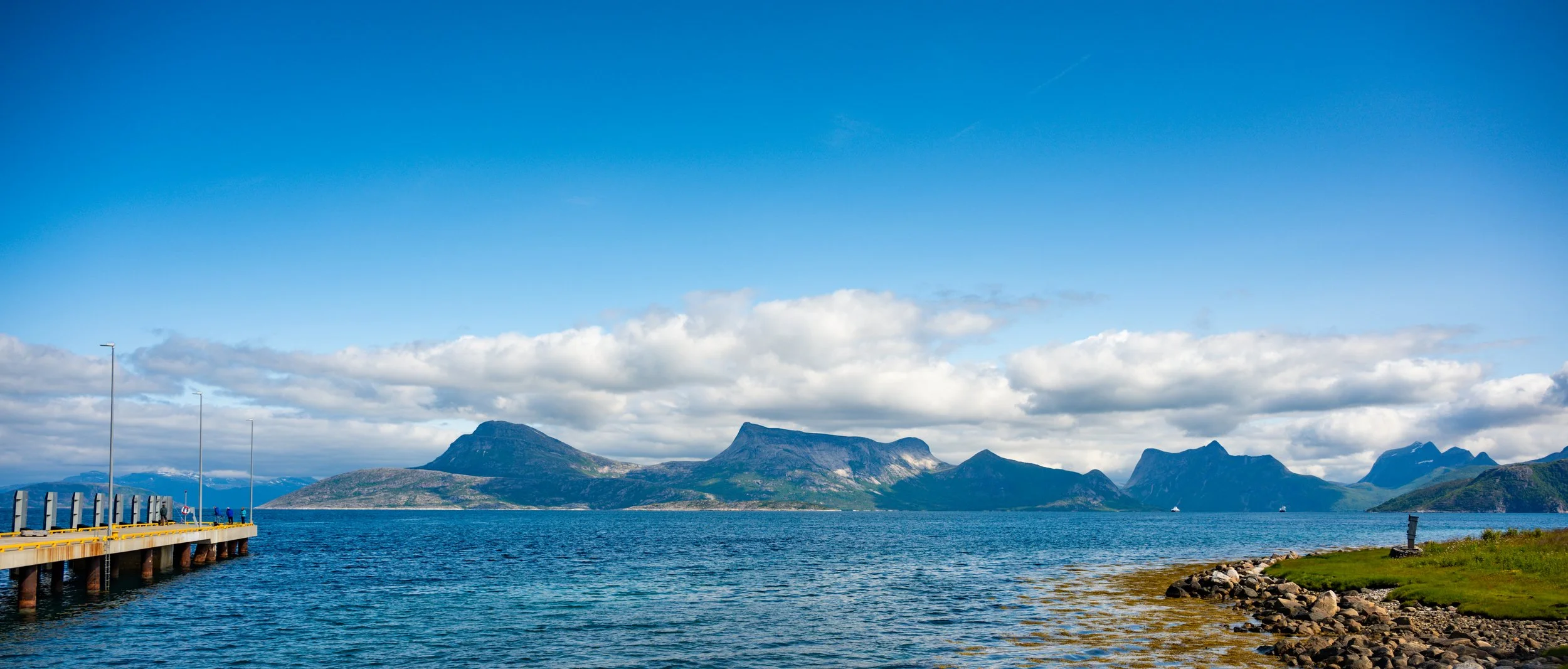 Scenic view of a lake with mountains in the background, a pier extending into the water on the left, and a rocky shoreline with green grass on the right under a partly cloudy sky.