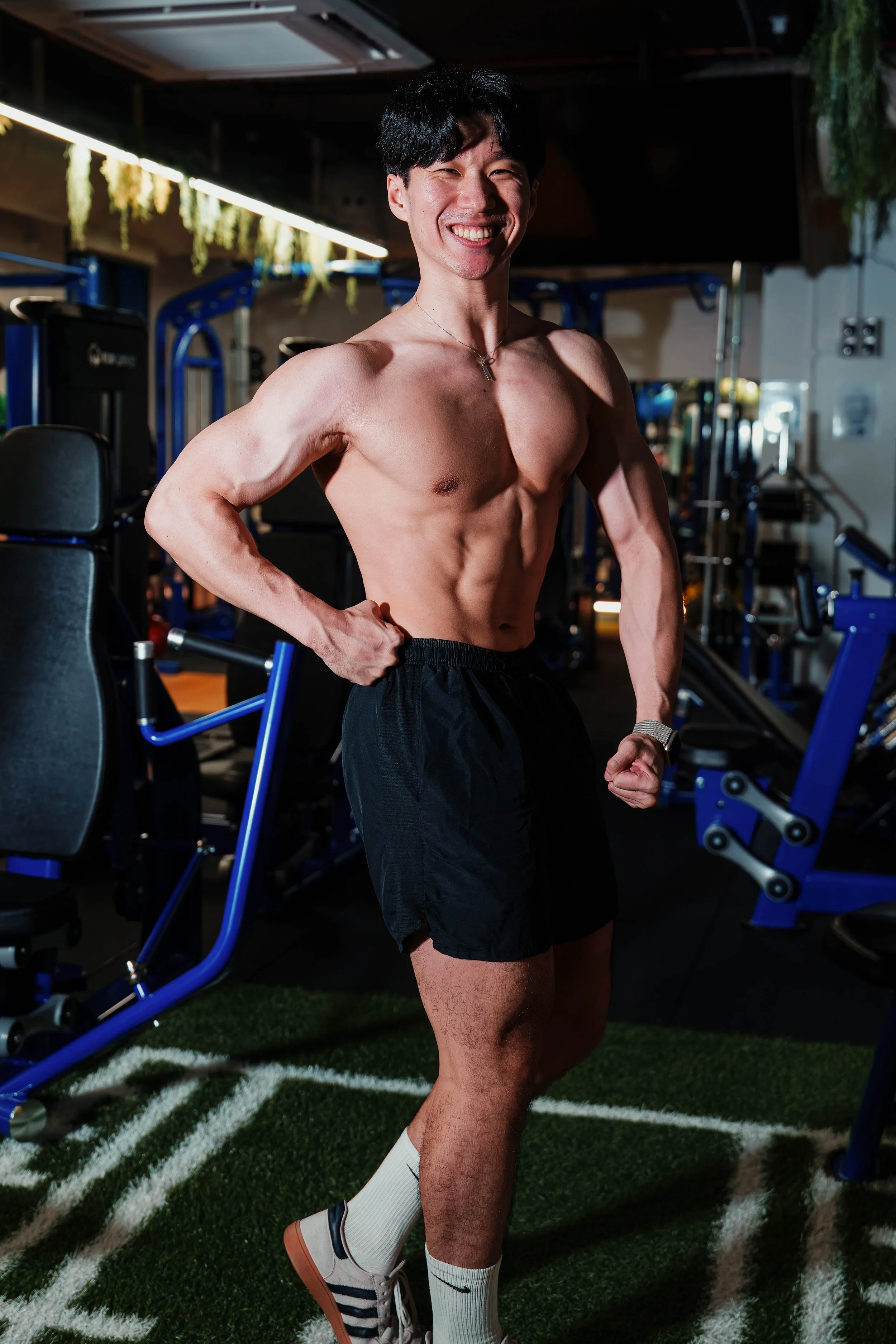 A shirtless man smiling and flexing his muscles in a gym, standing on artificial turf with gym equipment in the background.
