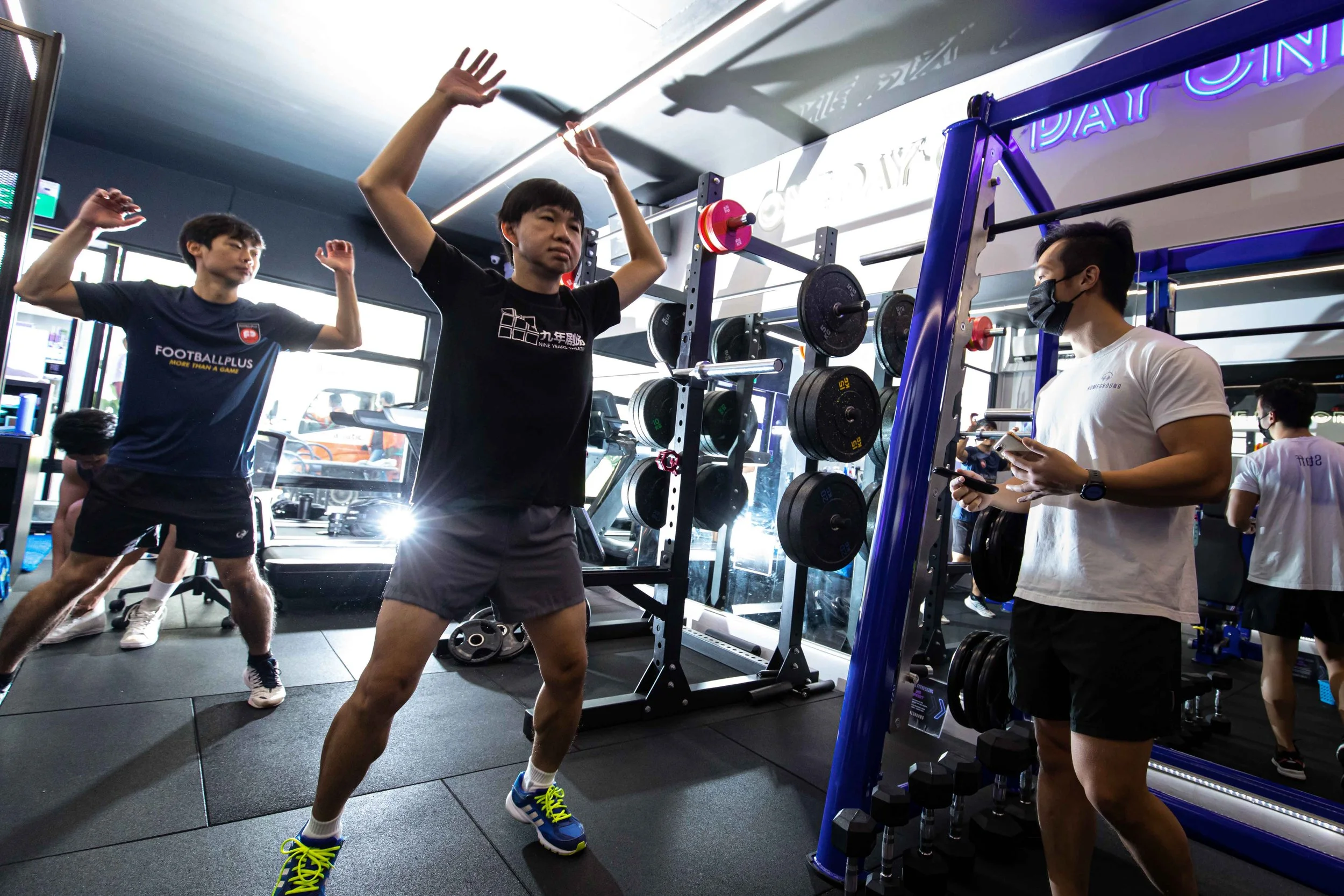 Three men in a gym, two doing exercises and one talking on a phone. The one in the center is stretching his arms upward, the one on the left is posing with arms bent, and the other man is wearing a white shirt and black mask, looking at his phone. Gym equipment and other people are visible in the background.