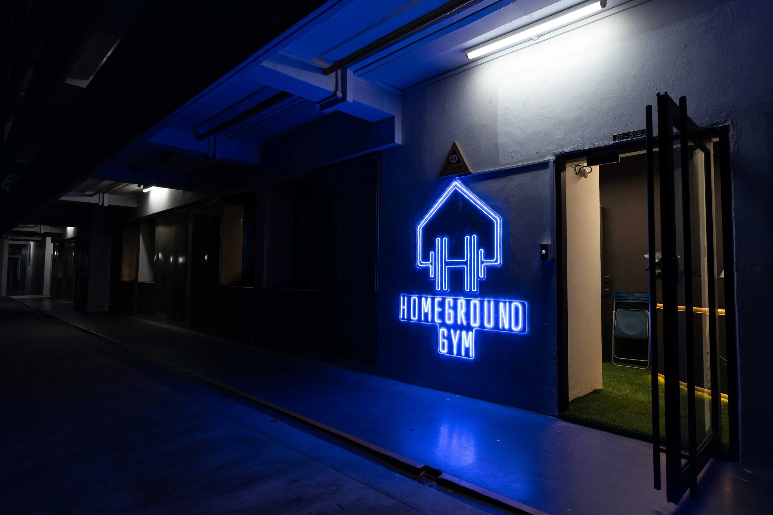Nighttime exterior view of a gym with a blue neon sign reading 'Homeground Gym' and an outline of a house with dumbbells inside. The entrance has a small grassy area and black metal gate, illuminated by a ceiling light.