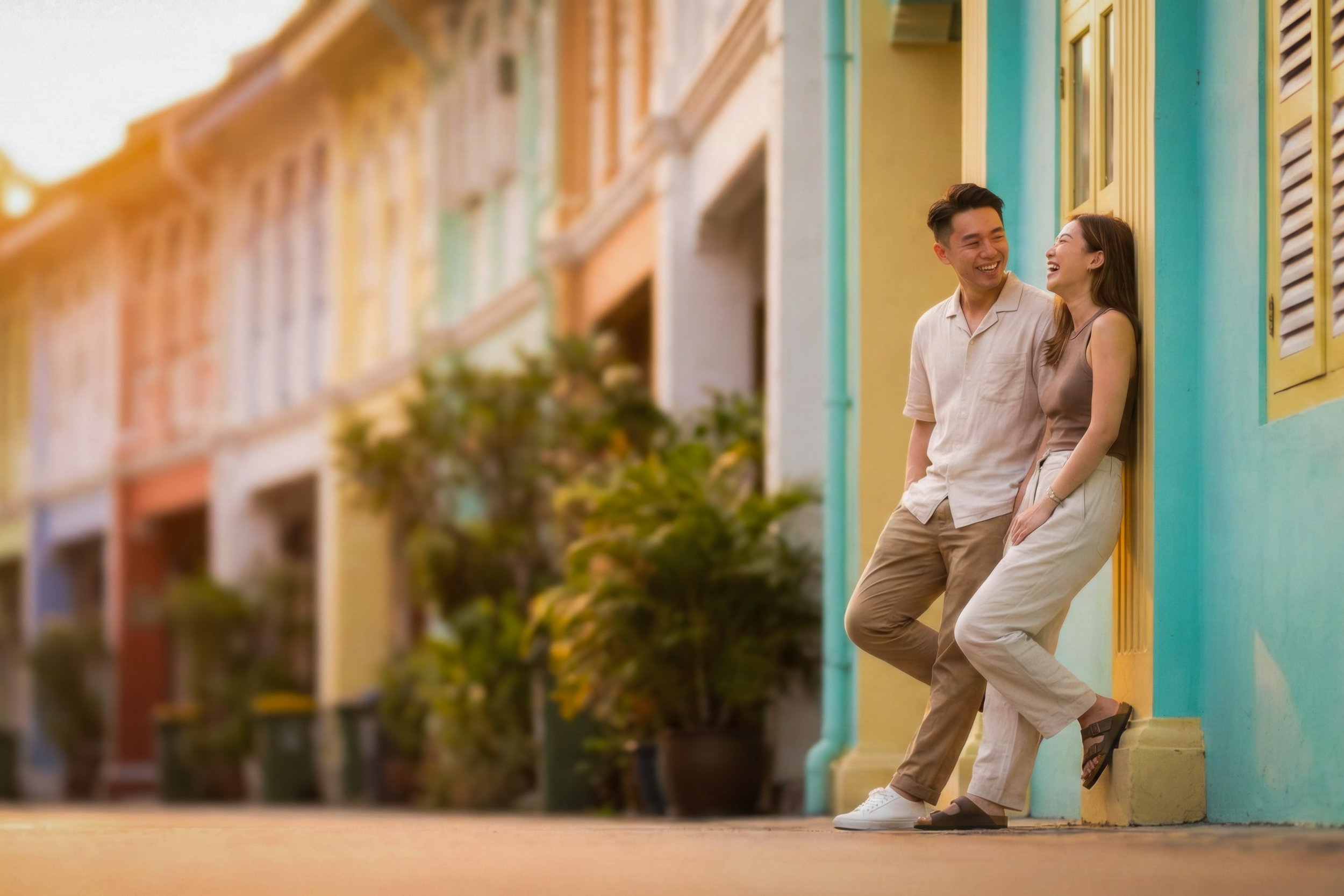 A young man and woman laughing and talking while leaning against colorful buildings on a street during sunset.