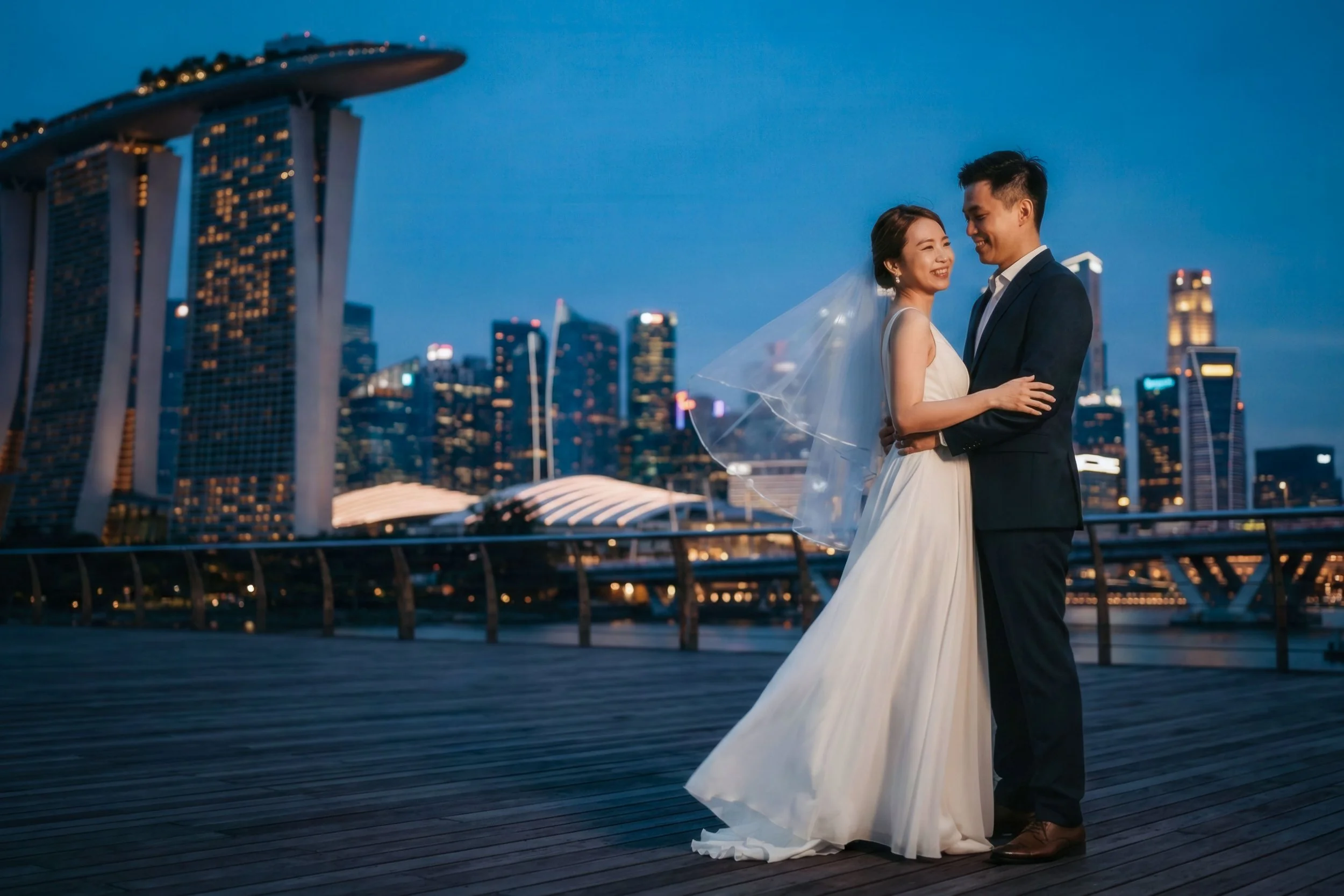 A bride and groom in wedding attire embracing on a waterfront promenade at dusk, with the Singapore skyline in the background, including the Marina Bay Sands hotel.