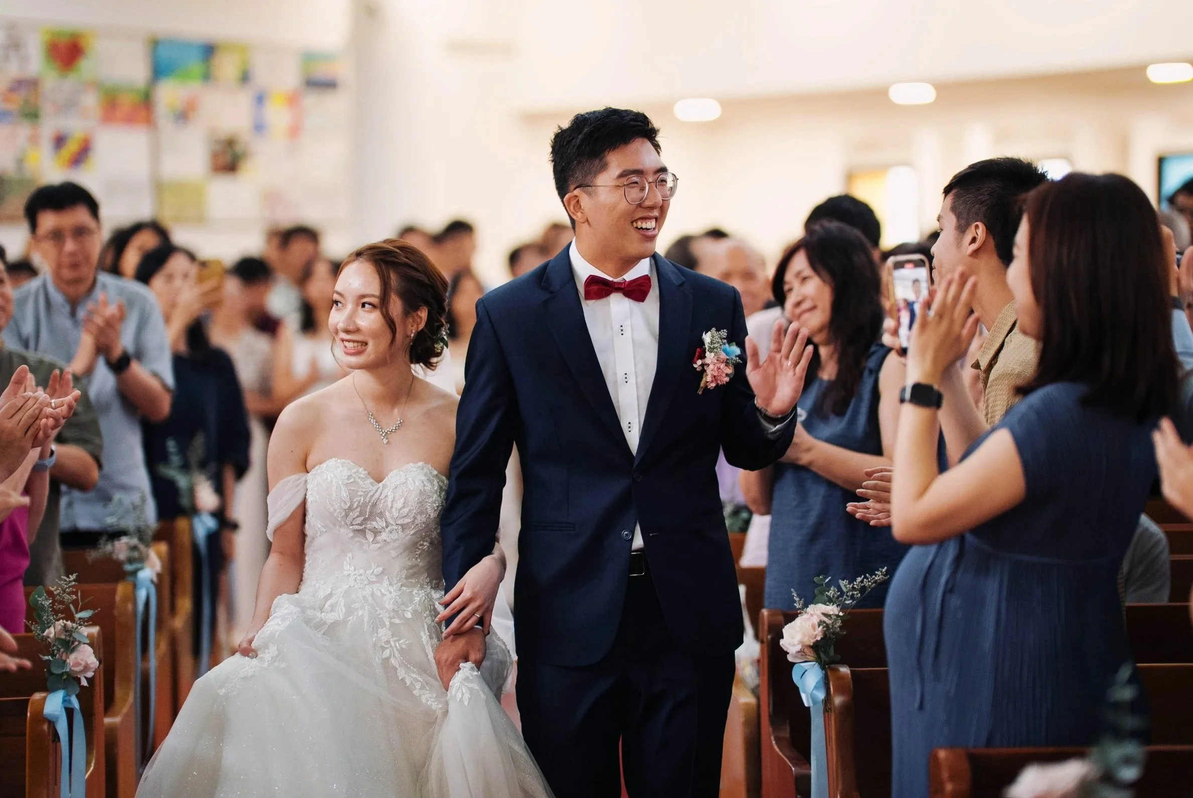 A bride and groom smiling and holding hands as they walk down the aisle during their wedding ceremony, surrounded by seated guests taking photos and clapping.