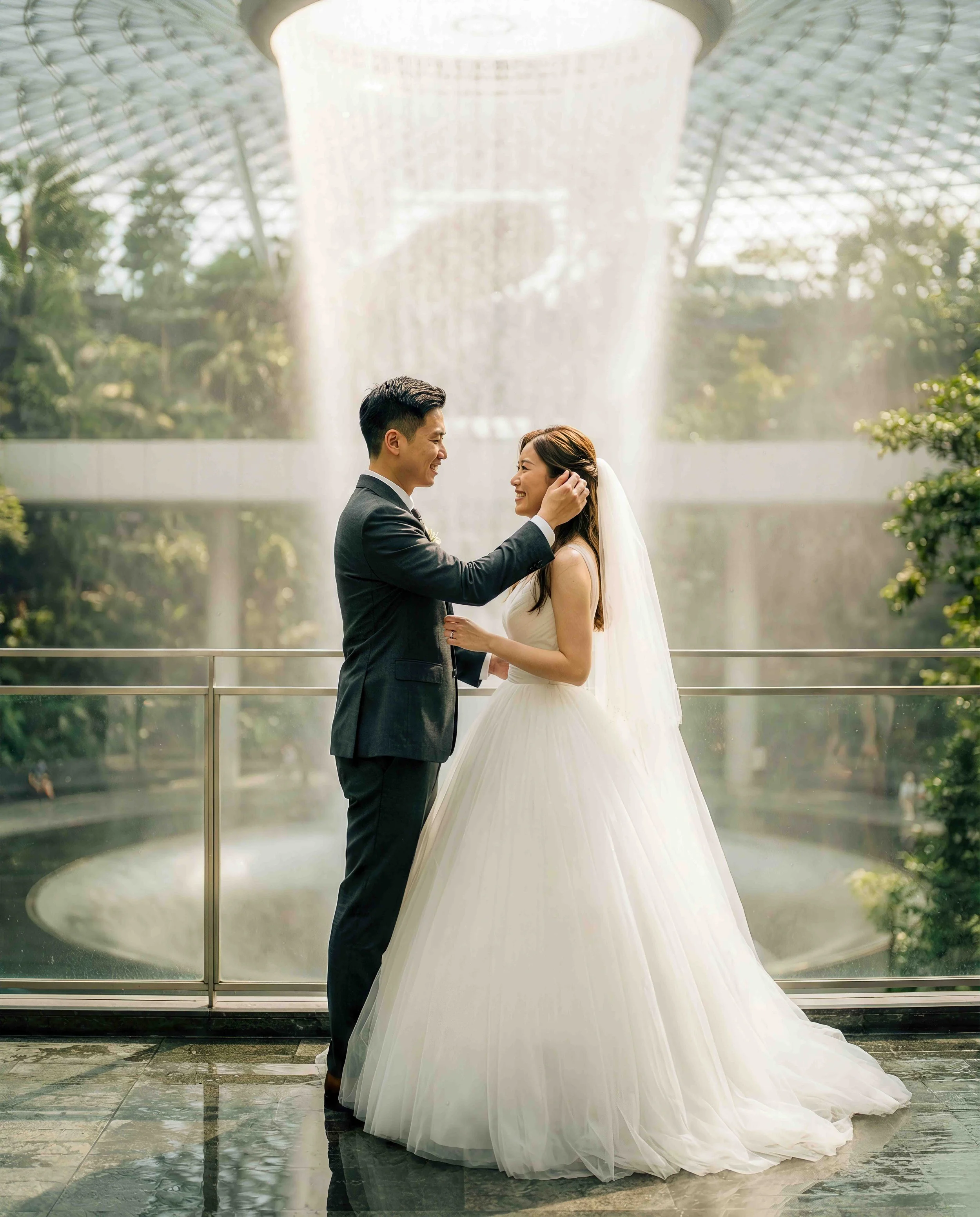 A bride in a white wedding gown and veil is smiling at a groom in a dark suit as he touches her face. They are standing in front of a large fountain with water cascading from above inside a modern glass and steel structure.
