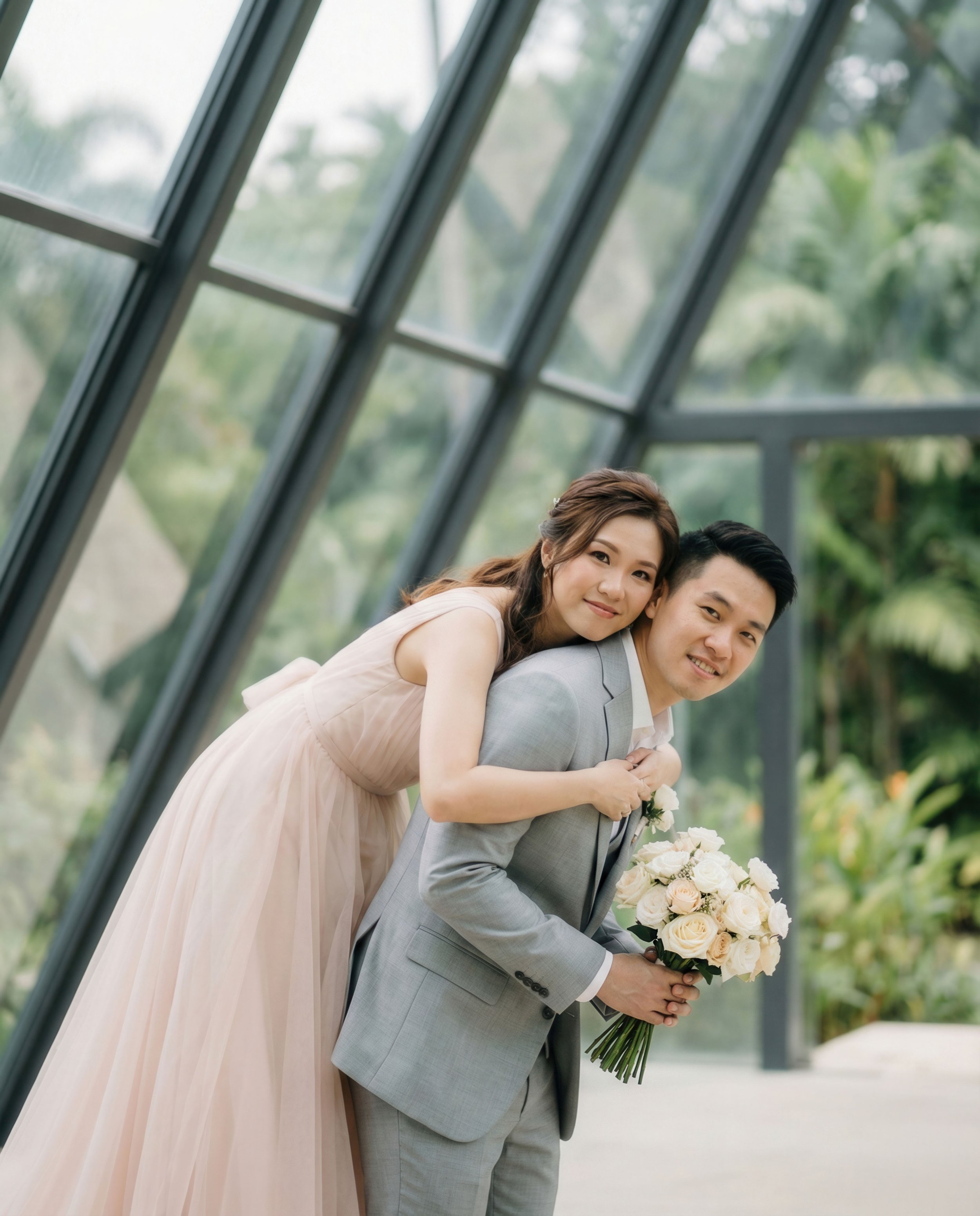 A couple on their wedding day, with the woman leaning on the man's back and holding a bouquet of white roses, inside a modern glass structure with greenery outside.