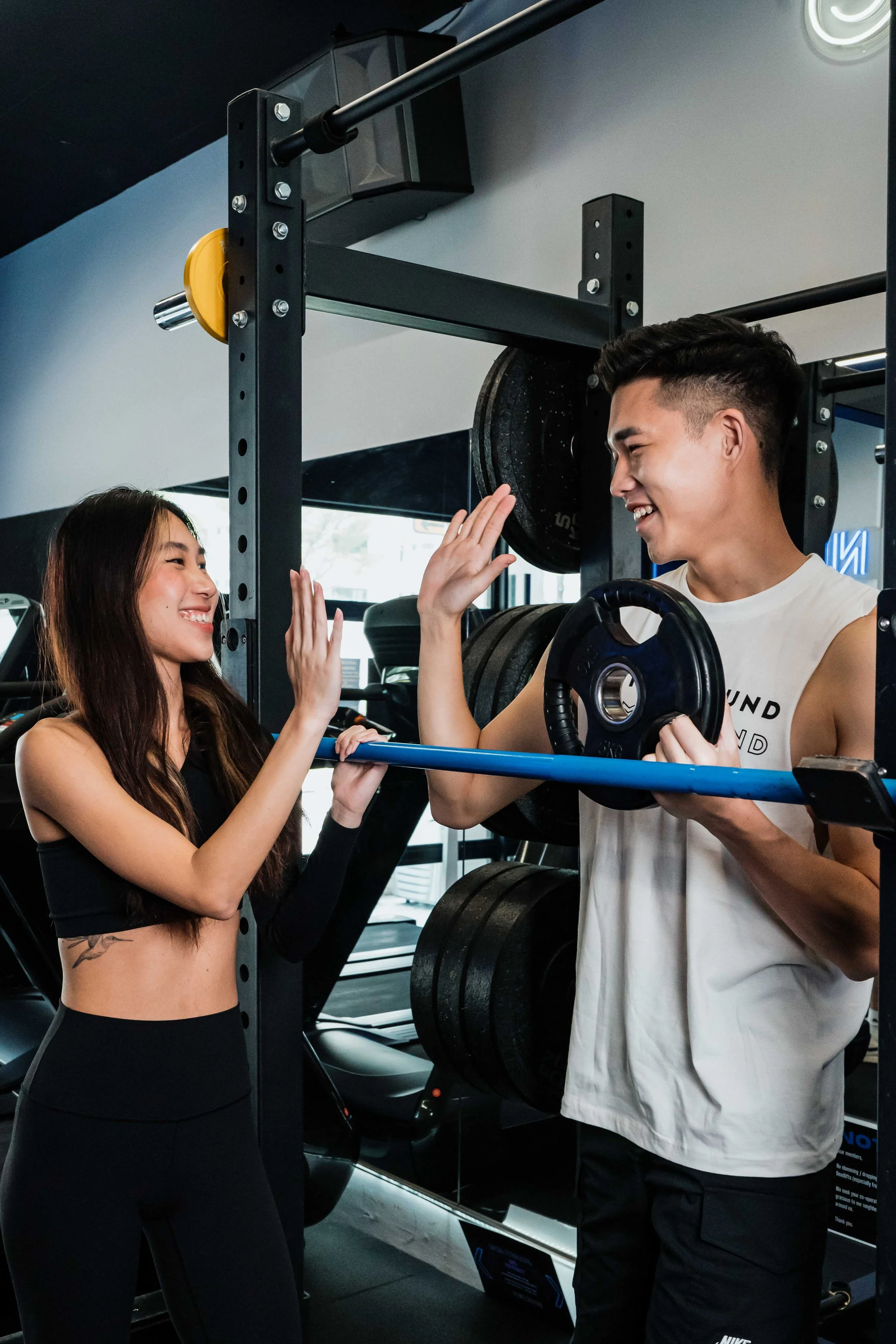 A young woman and a young man high-fiving in a gym, with the woman holding a barbell and the man holding a weight plate, both smiling.