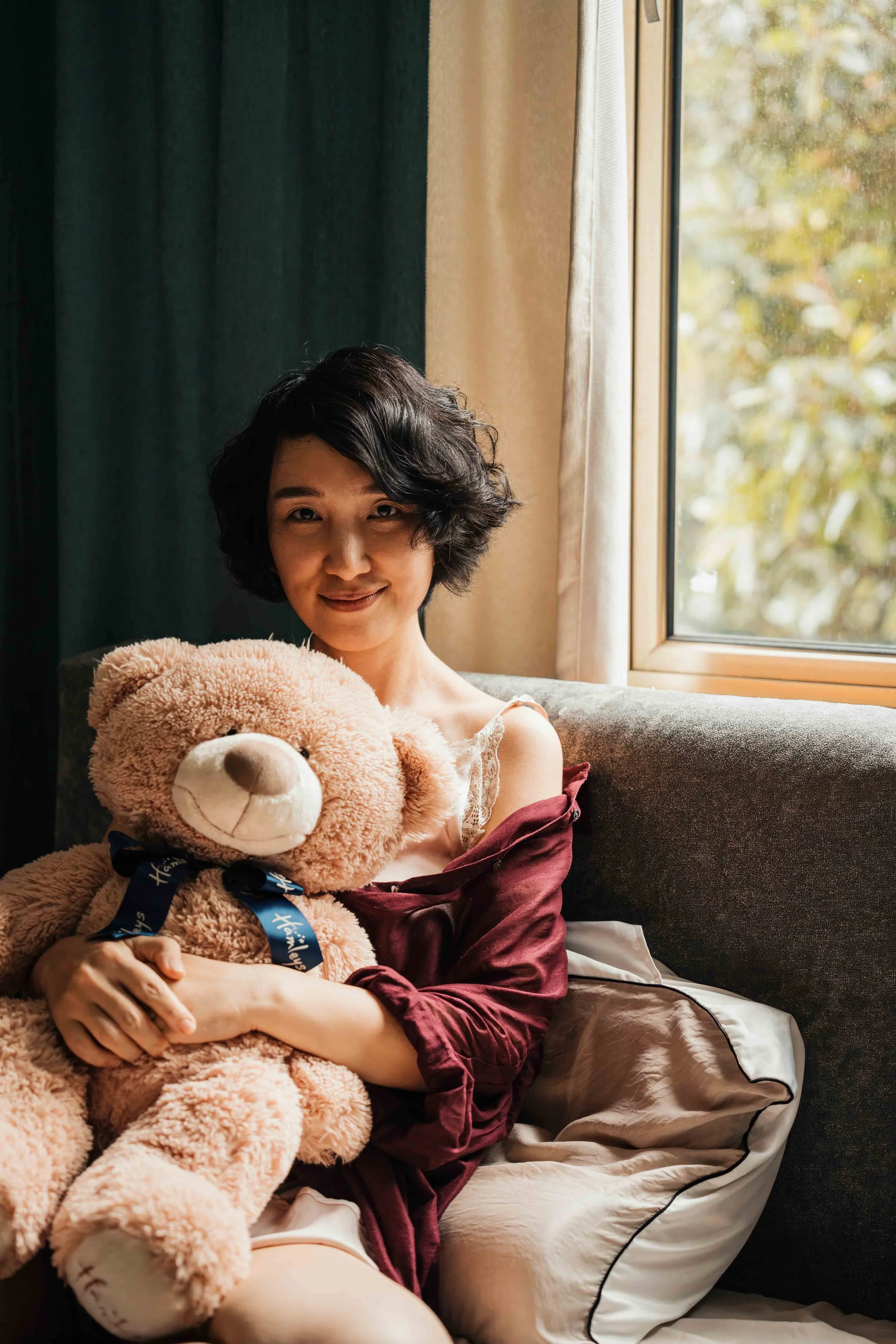 A woman with short black hair sitting on a couch, holding a large teddy bear, near a window with beige and green curtains, with trees visible outside.