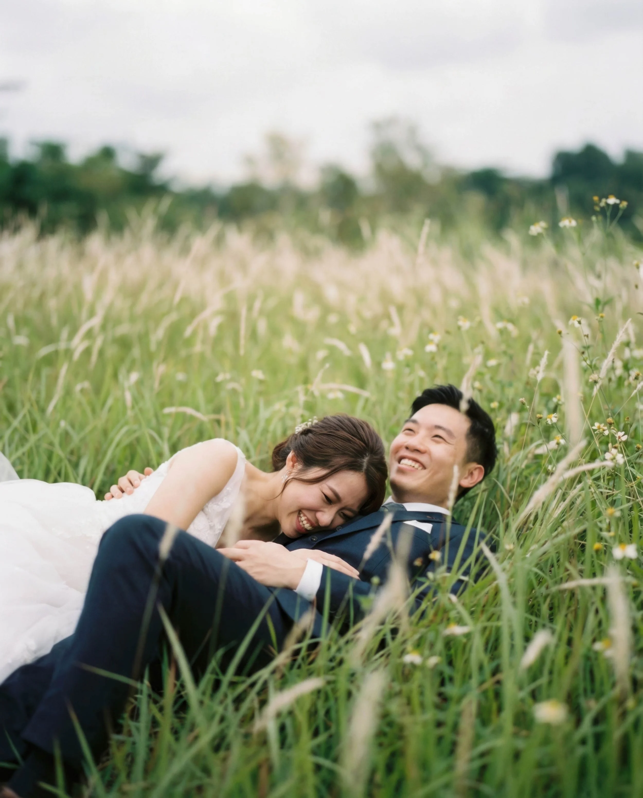 A happy couple in wedding attire lying in a grassy field, smiling and laughing.