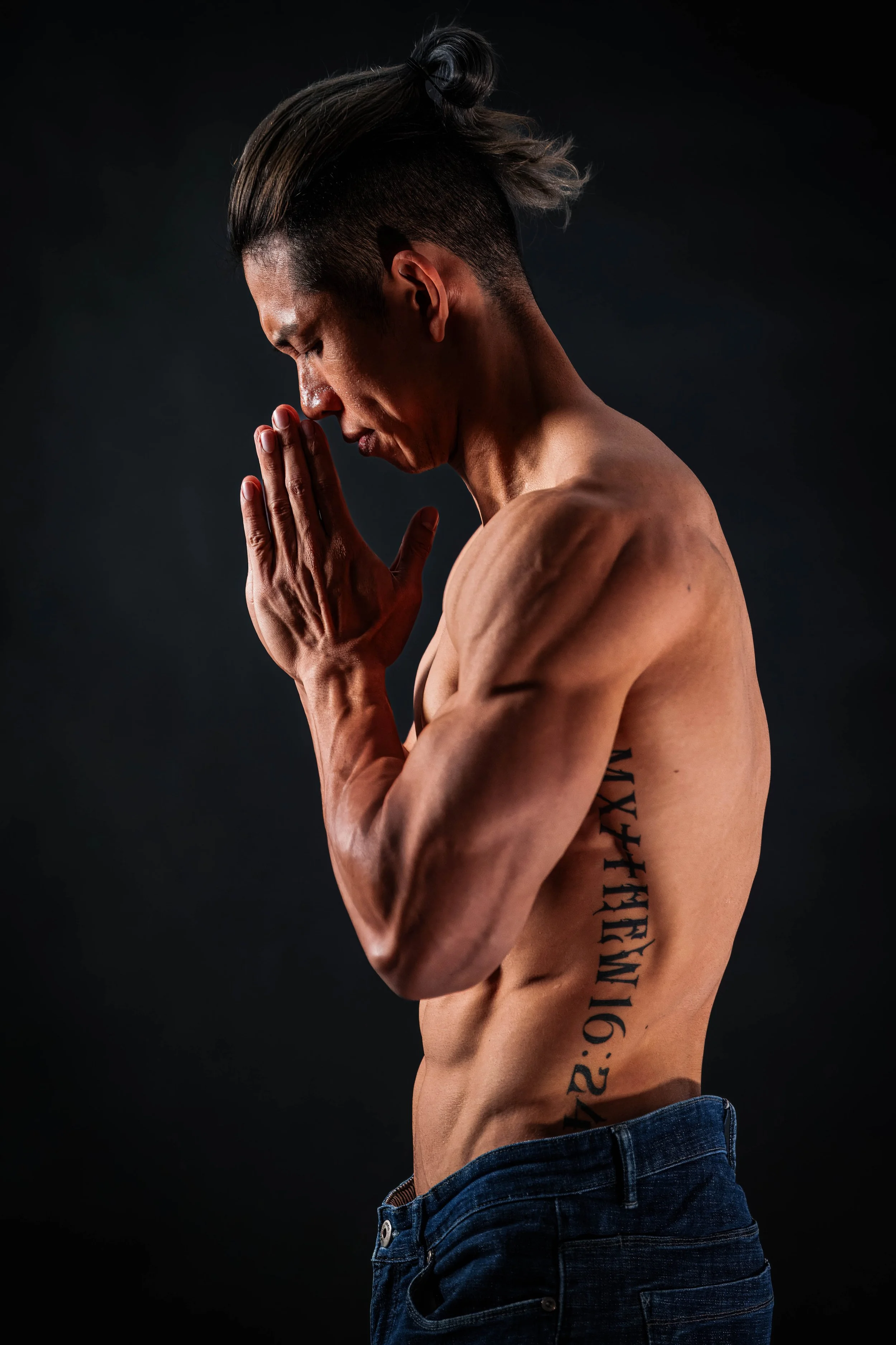 A shirtless man with a tattoo on his side stands with hands pressed together in prayer or meditation, eyes closed, against a dark background.