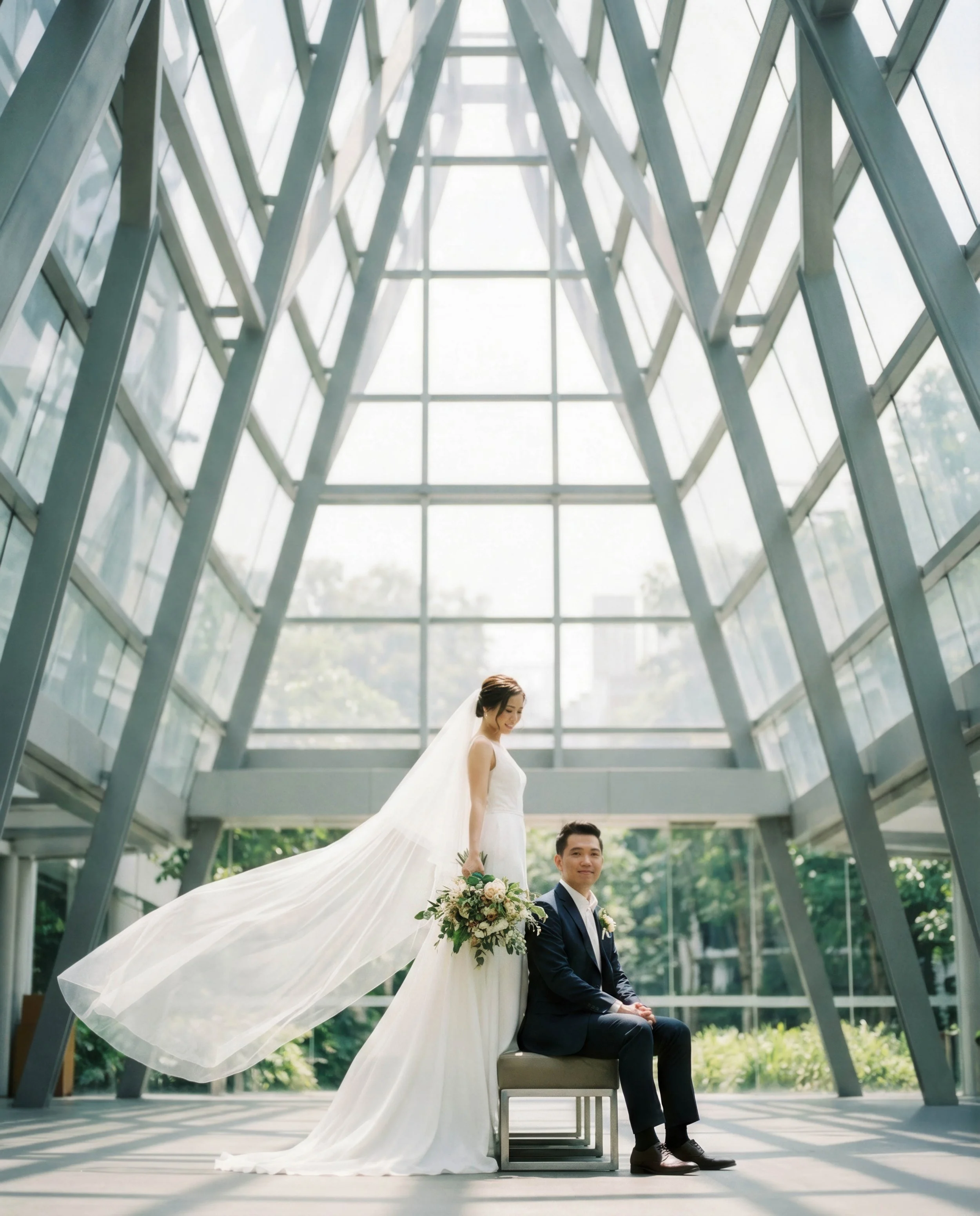 A bride and groom in a modern glass-walled building with sunlight streaming through the large windows. The bride is standing behind the seated groom, holding a bouquet of flowers, with her veil flowing behind her.