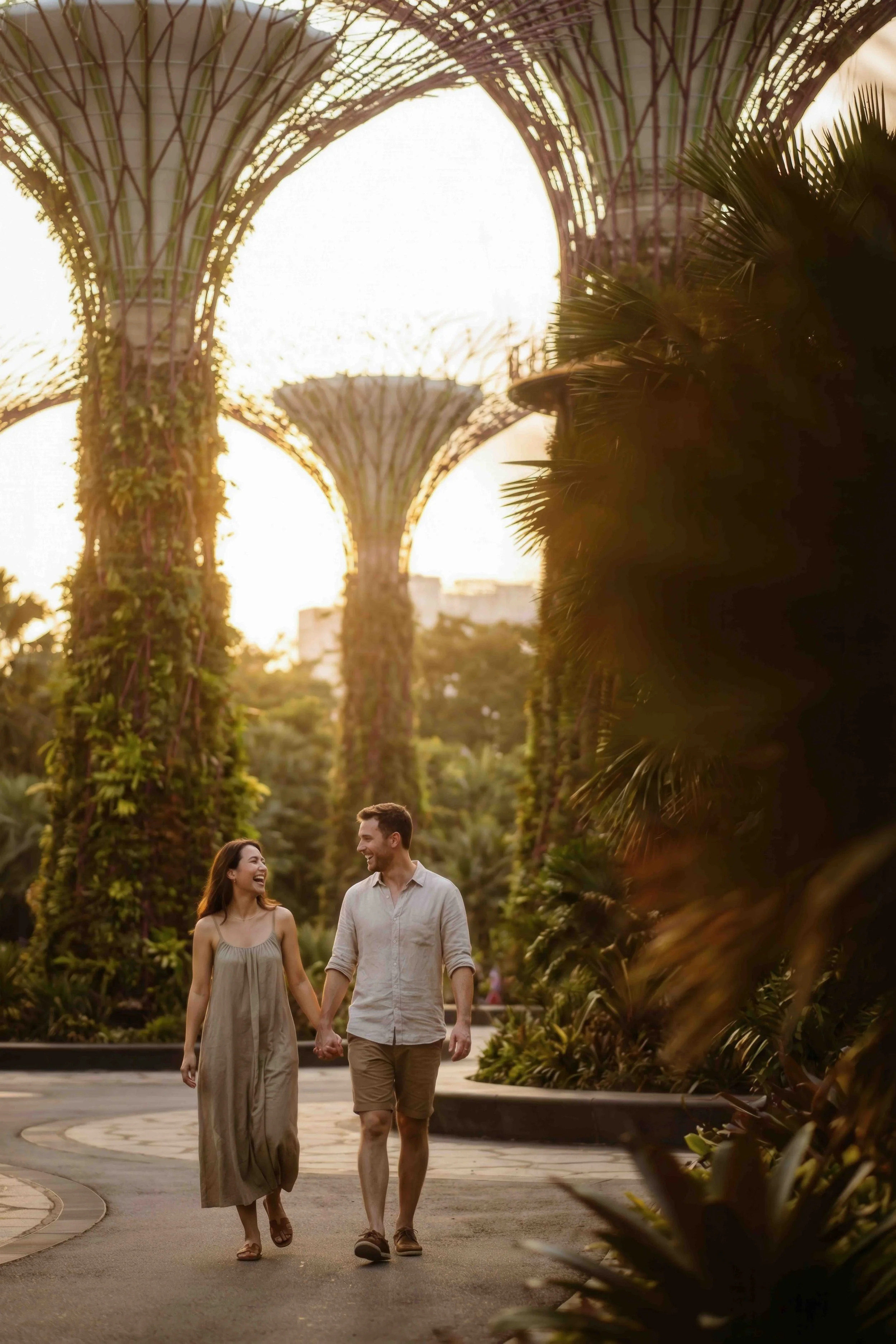 A young couple is walking hand-in-hand through a lush, green garden during sunset, smiling at each other.