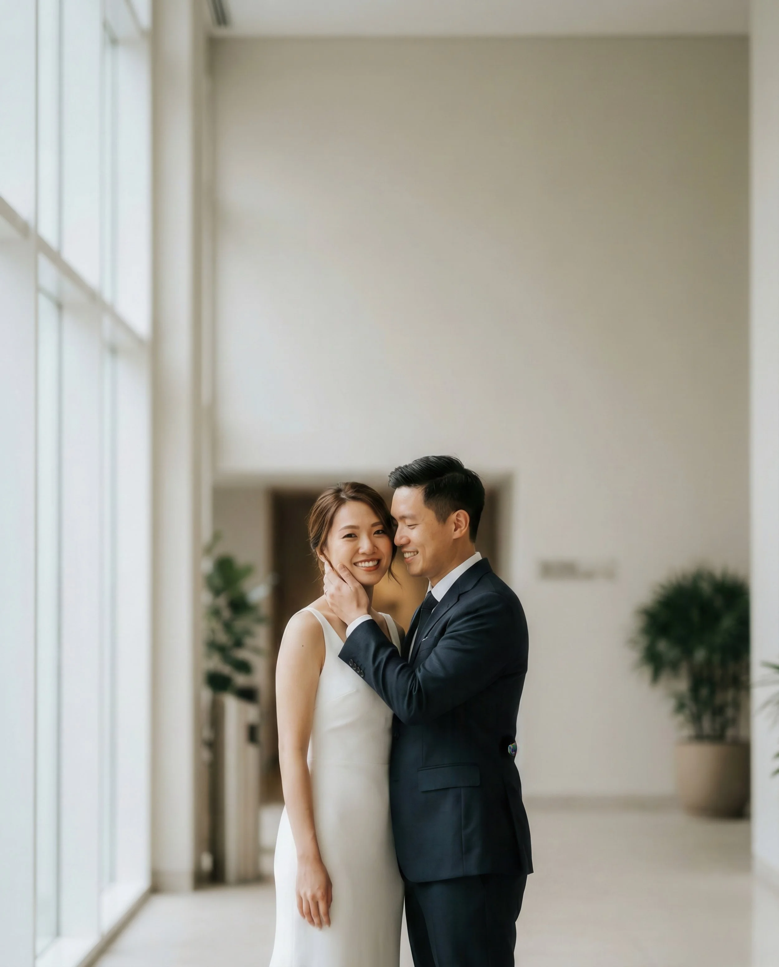 A happy couple in wedding attire smiling and embracing indoors, with large windows and potted plants in the background.