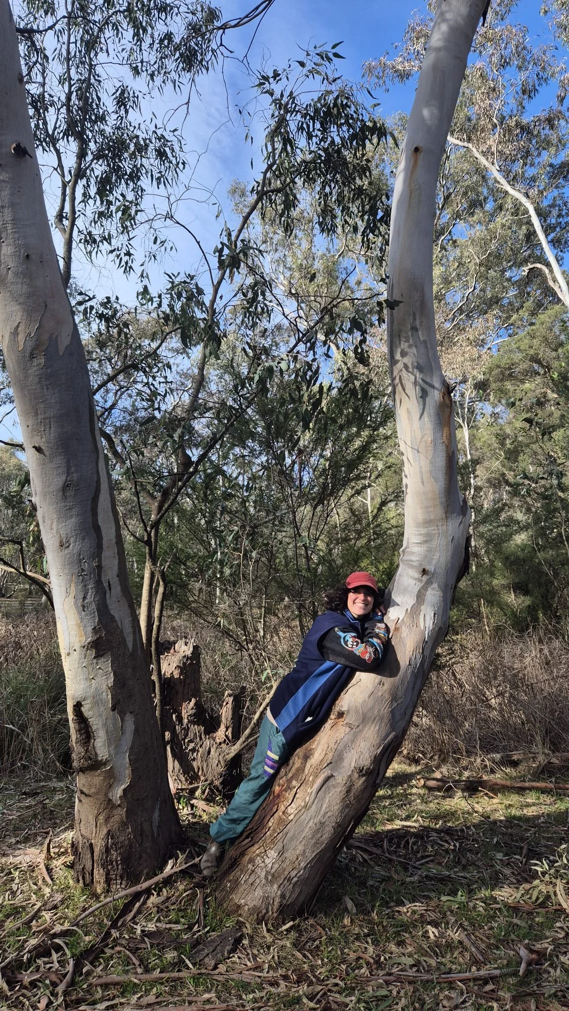 A smiling woman in outdoor clothing leaning on a tree trunk in a wooded area on a sunny day.