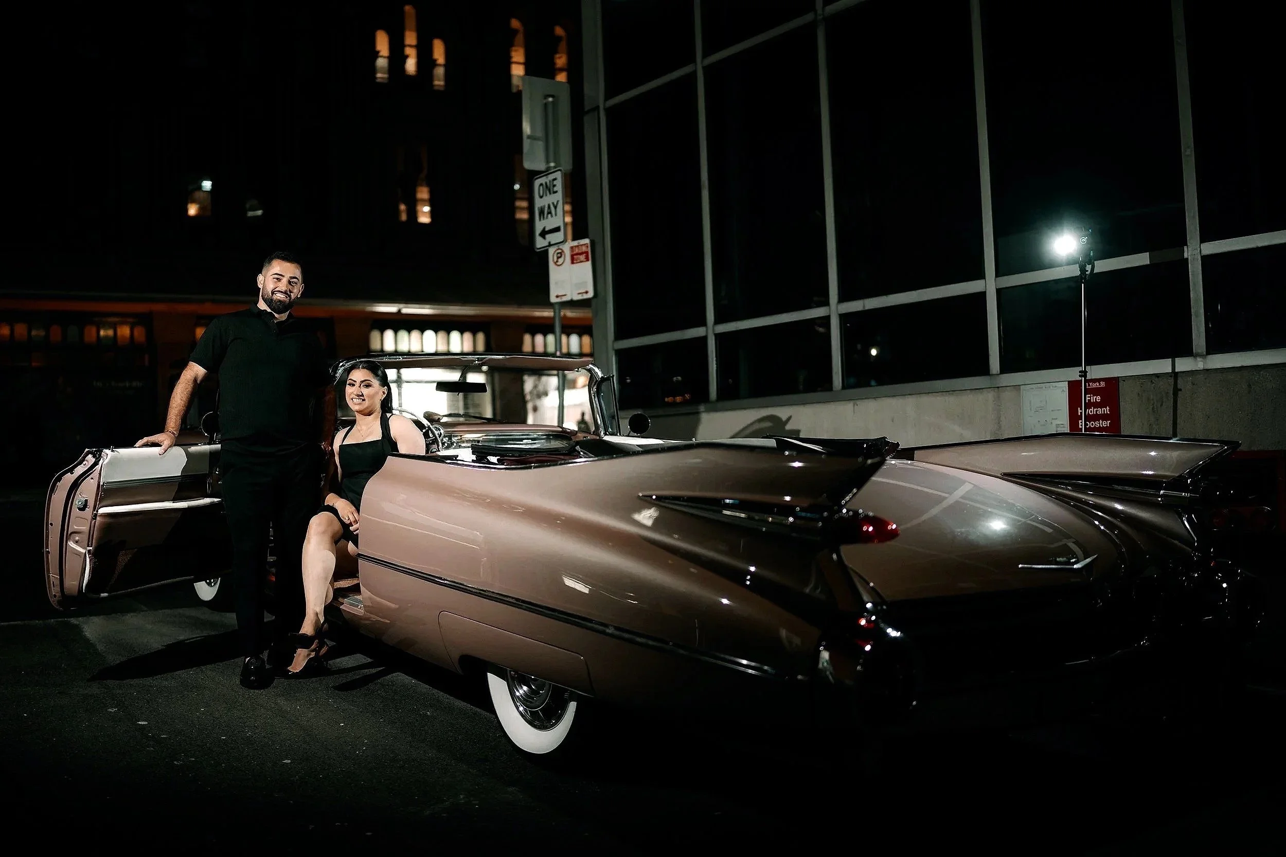 A man and a woman in formal attire posing with a vintage Cadillac car at night in an urban setting.