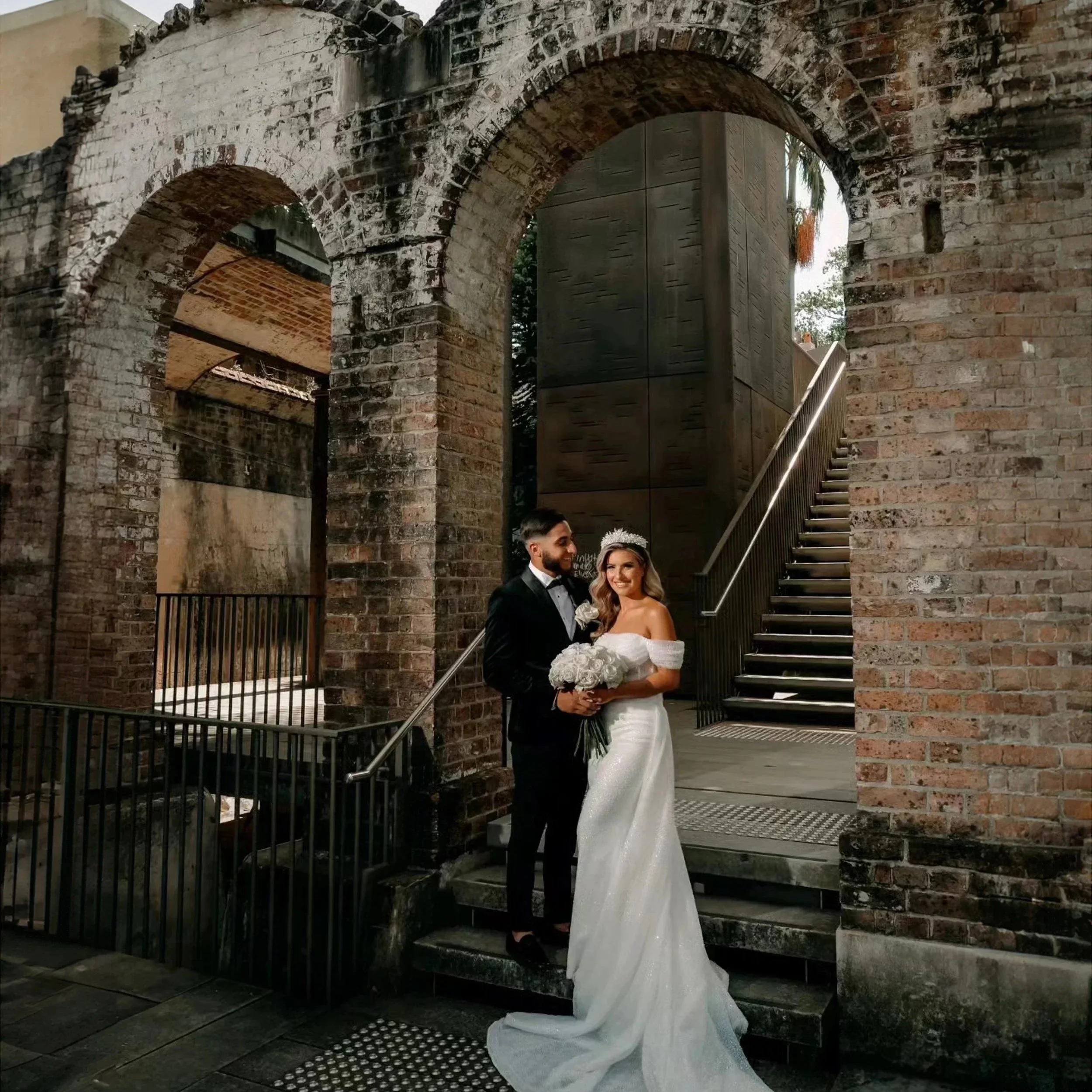 Bride and groom standing on steps beneath a brick archway, with the bride holding a bouquet of white flowers.