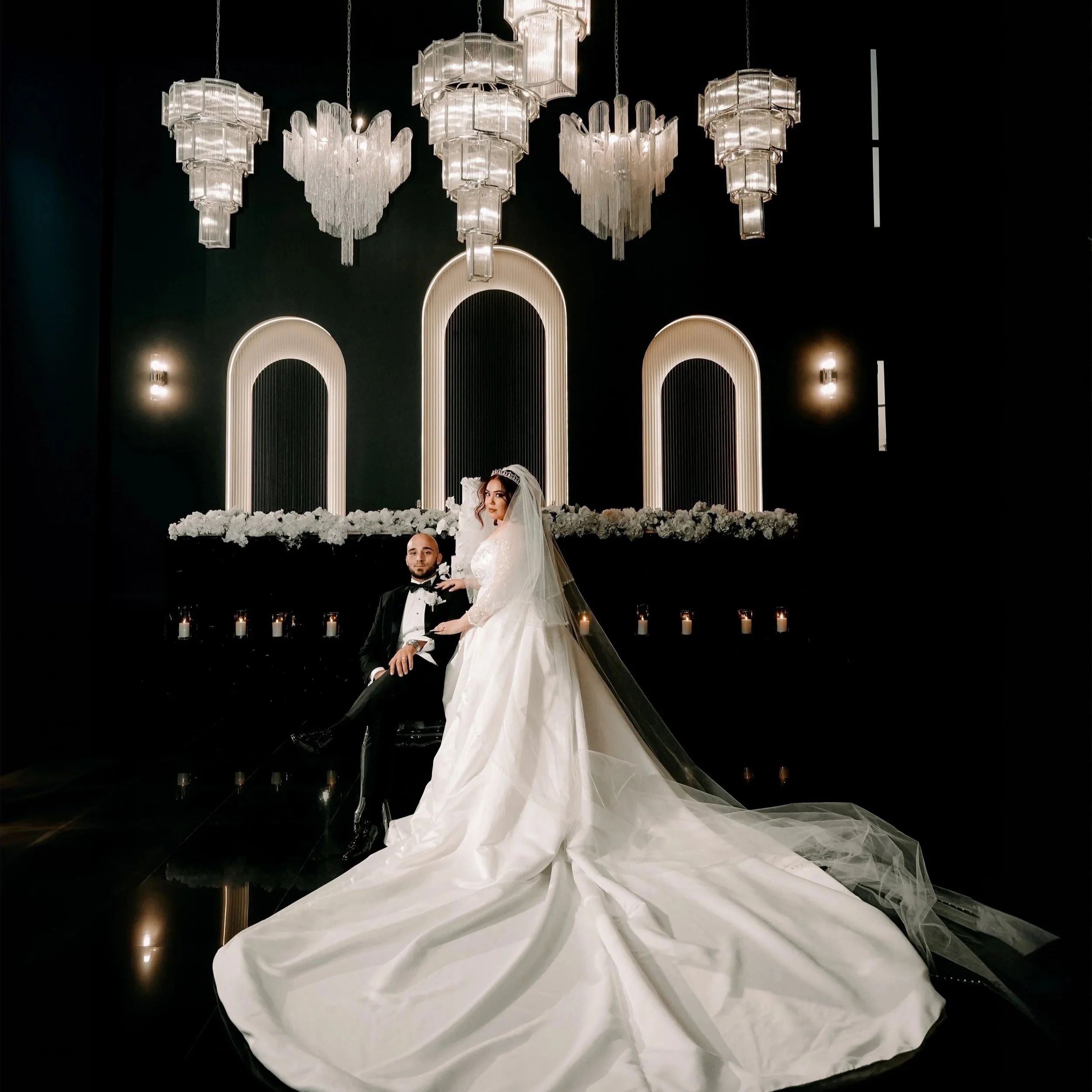A bride and groom pose together in a modern, elegant wedding venue with a black background, stylish lighting, and floral decorations.