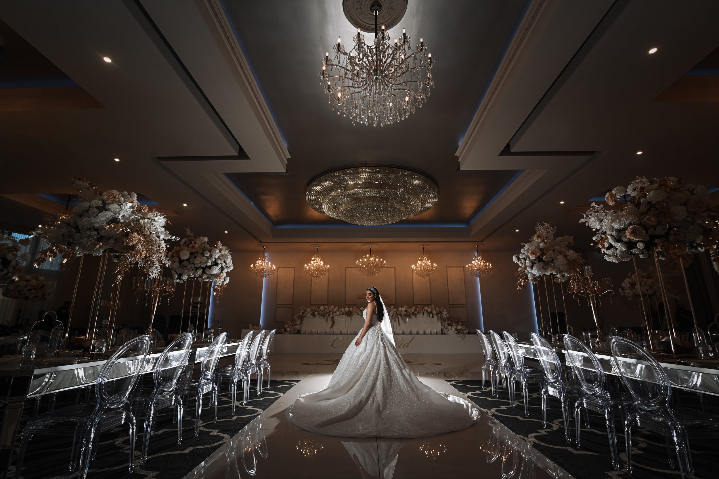 Bride in wedding gown standing in an elegant decorated banquet hall with large chandeliers and floral centerpieces.