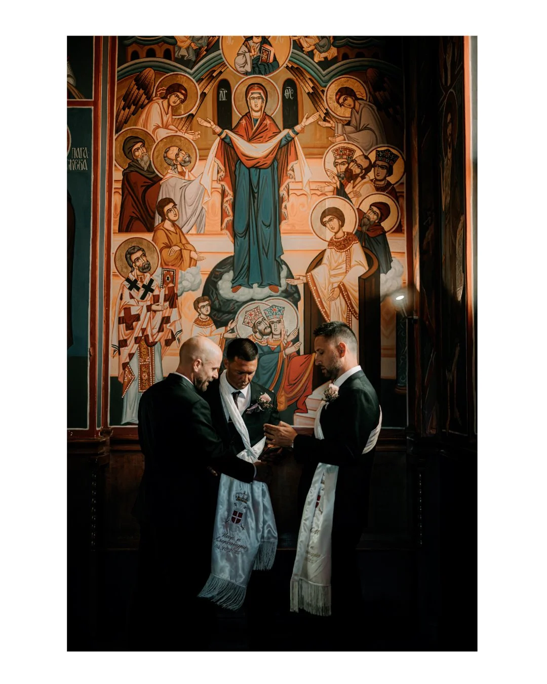Three men in suits engaged in a religious ceremony in front of a large religious icon painting depicting the Virgin Mary, Jesus, and angels, inside a church.