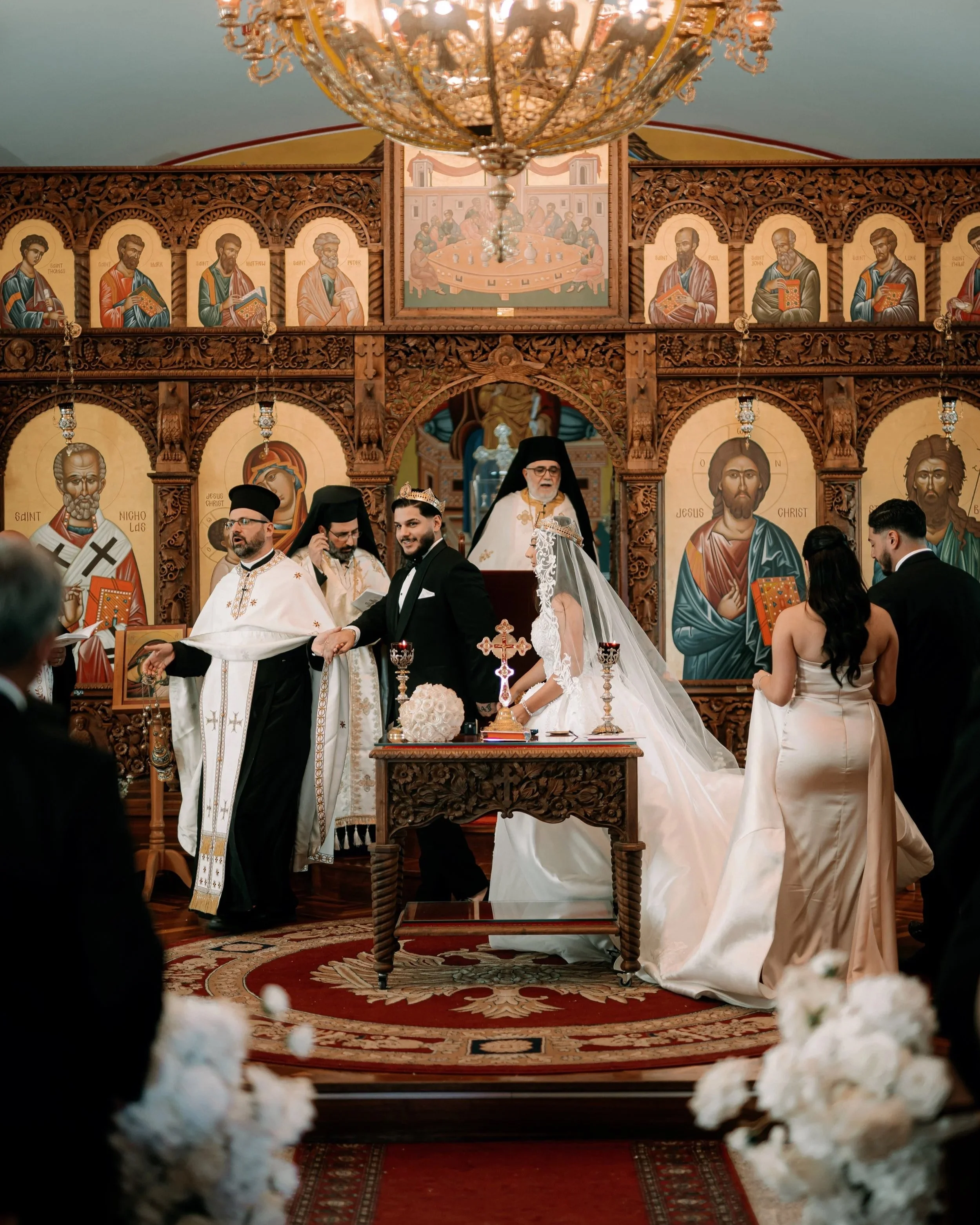 A wedding ceremony taking place in an St Nicholas Antiochian Orthodox Church Punchbowl, with a priest, bride, groom, and wedding party, surrounded by religious iconography and ornate wooden decor.