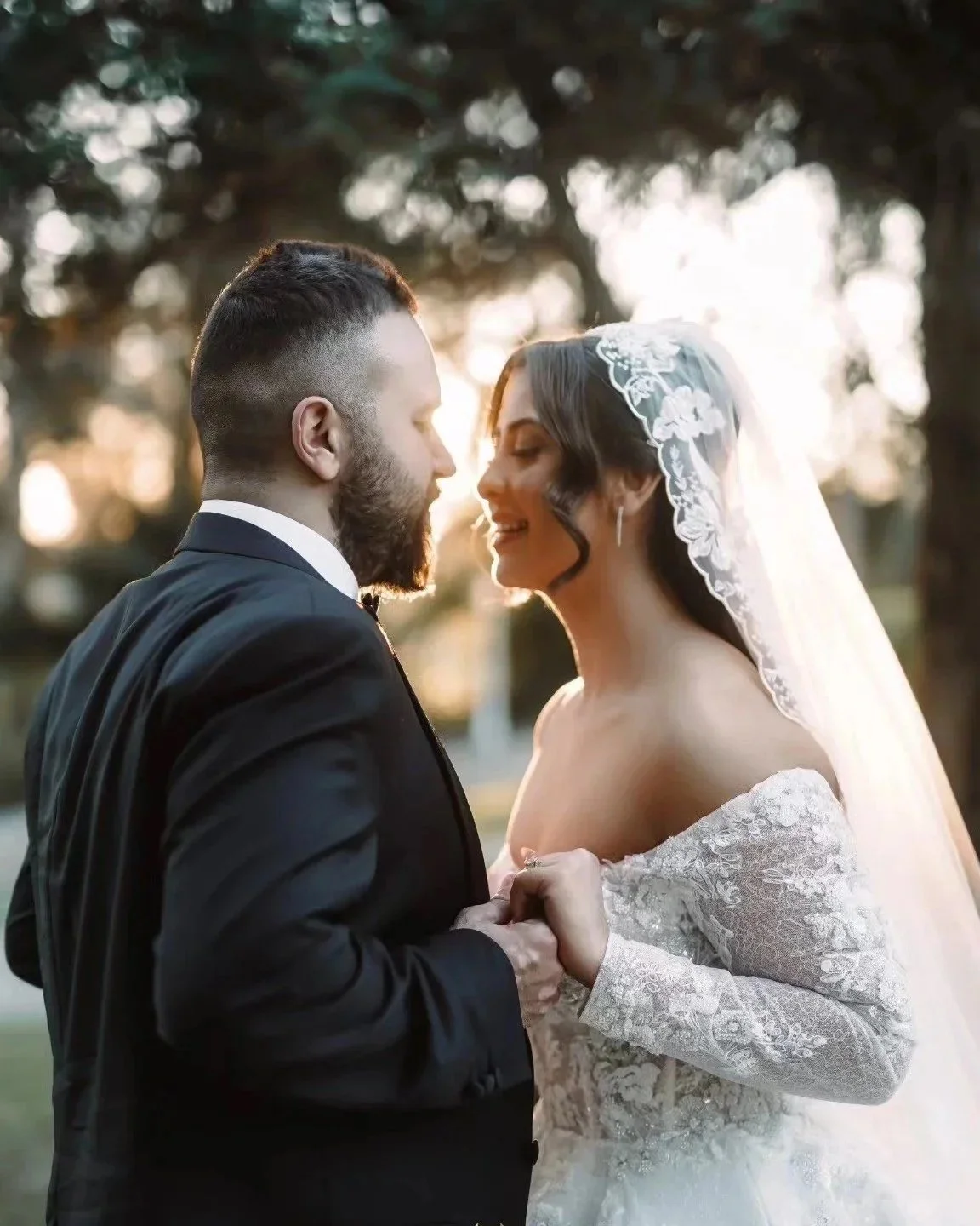 A bride and groom holding hands and smiling at each other outdoors during sunset, with trees in the background.