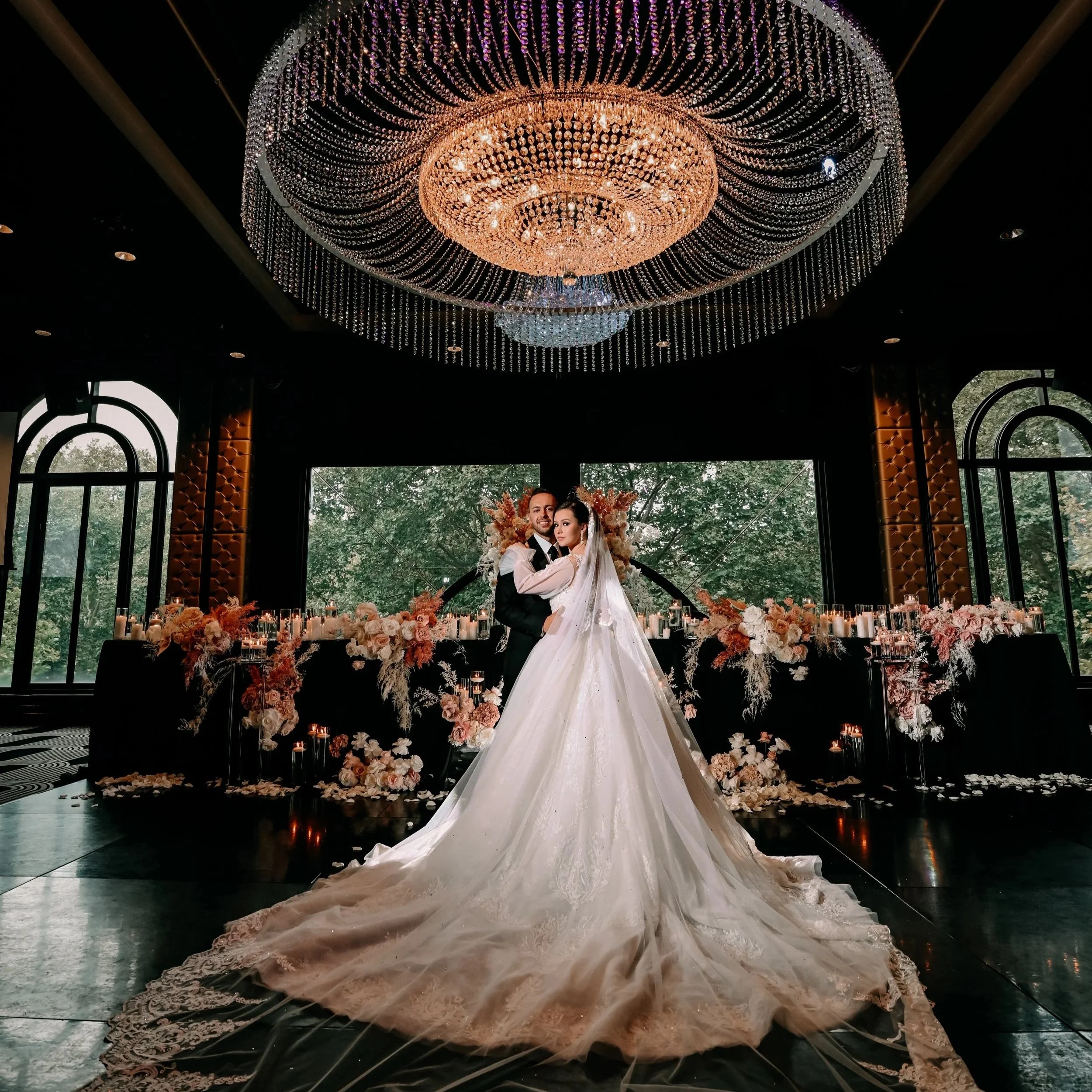 A bride and groom pose together in wedding attire inside a luxurious venue with a large, ornate chandelier overhead and large windows showing trees outside. The bride wears a white, long-sleeved wedding gown with a full skirt and a veil, while the groom wears a black tuxedo. The background features a table decorated with pink and white flowers and candles.