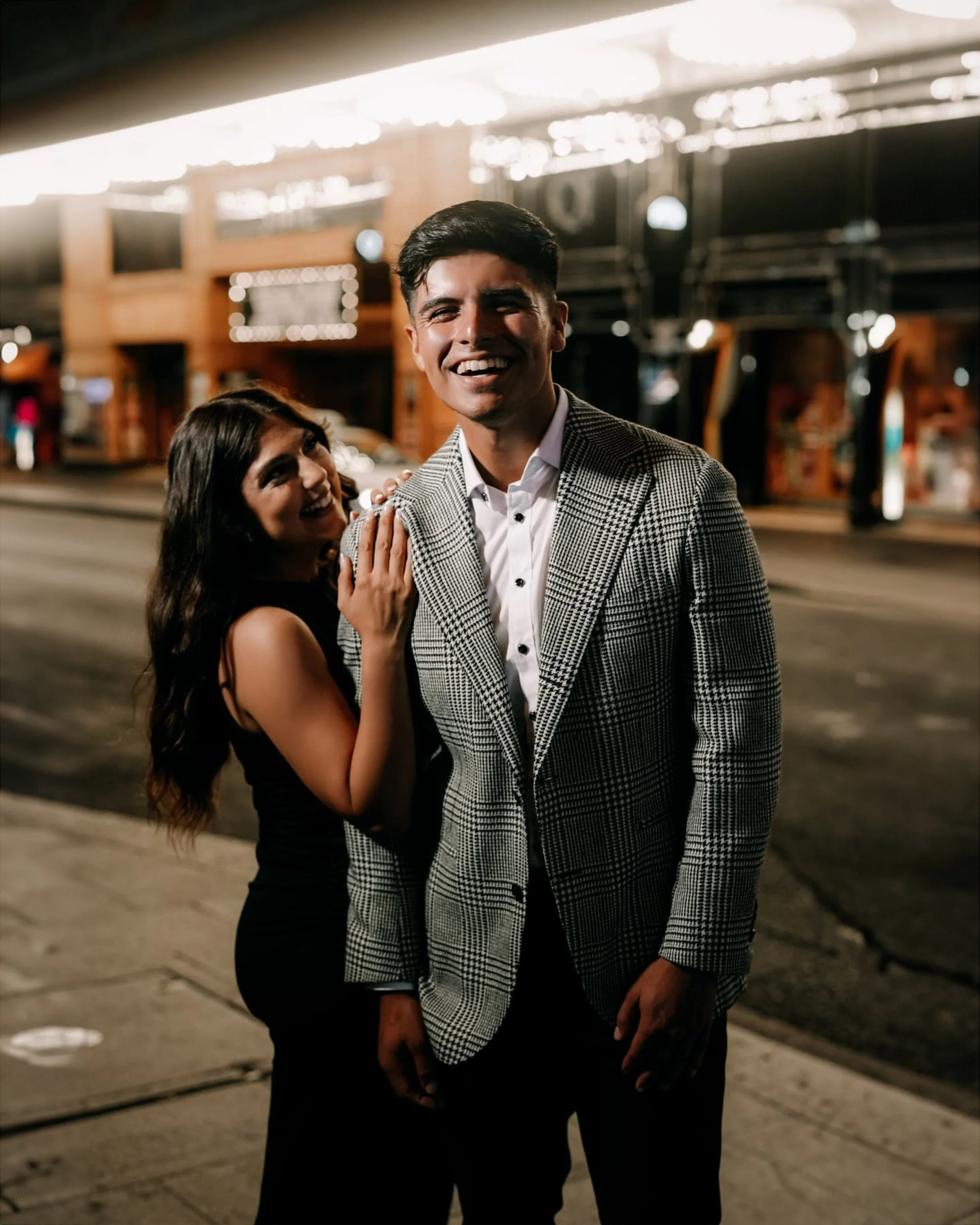 A young couple standing on a city sidewalk at night, smiling and laughing. The man is wearing a checkered blazer and white shirt, and the woman is in a black sleeveless dress and has her hand on his shoulder.