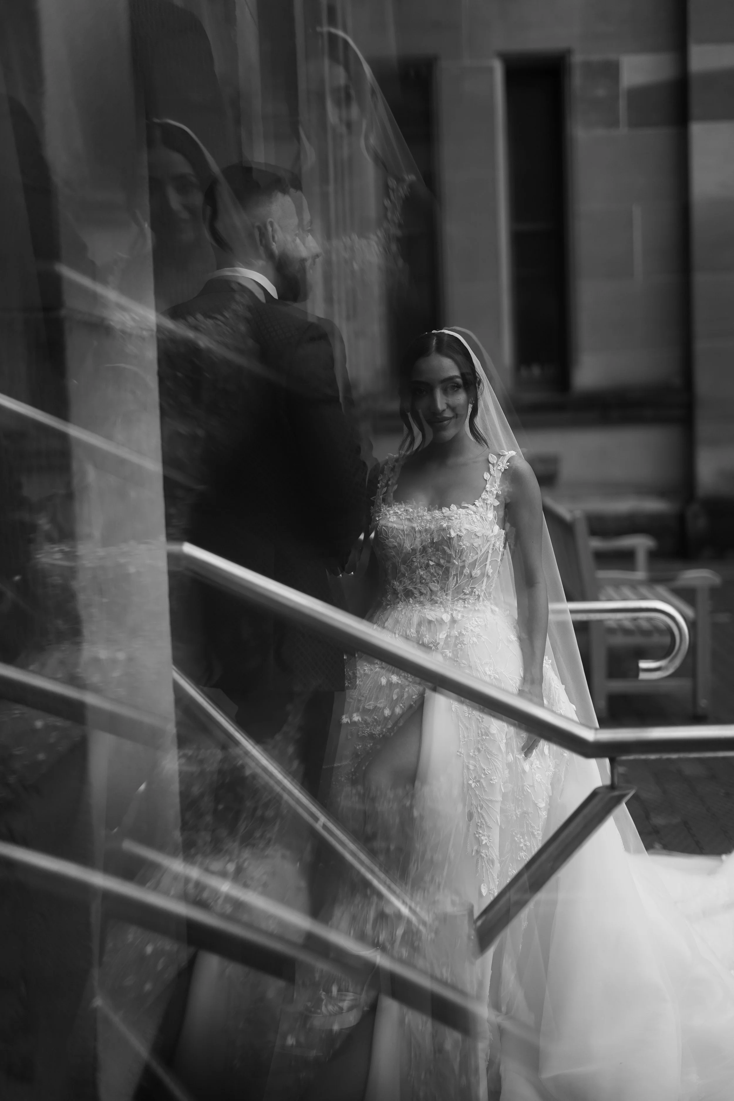 Bride in a wedding gown and veil smiling, seen through a glass window with reflections, with a man in a suit behind her.