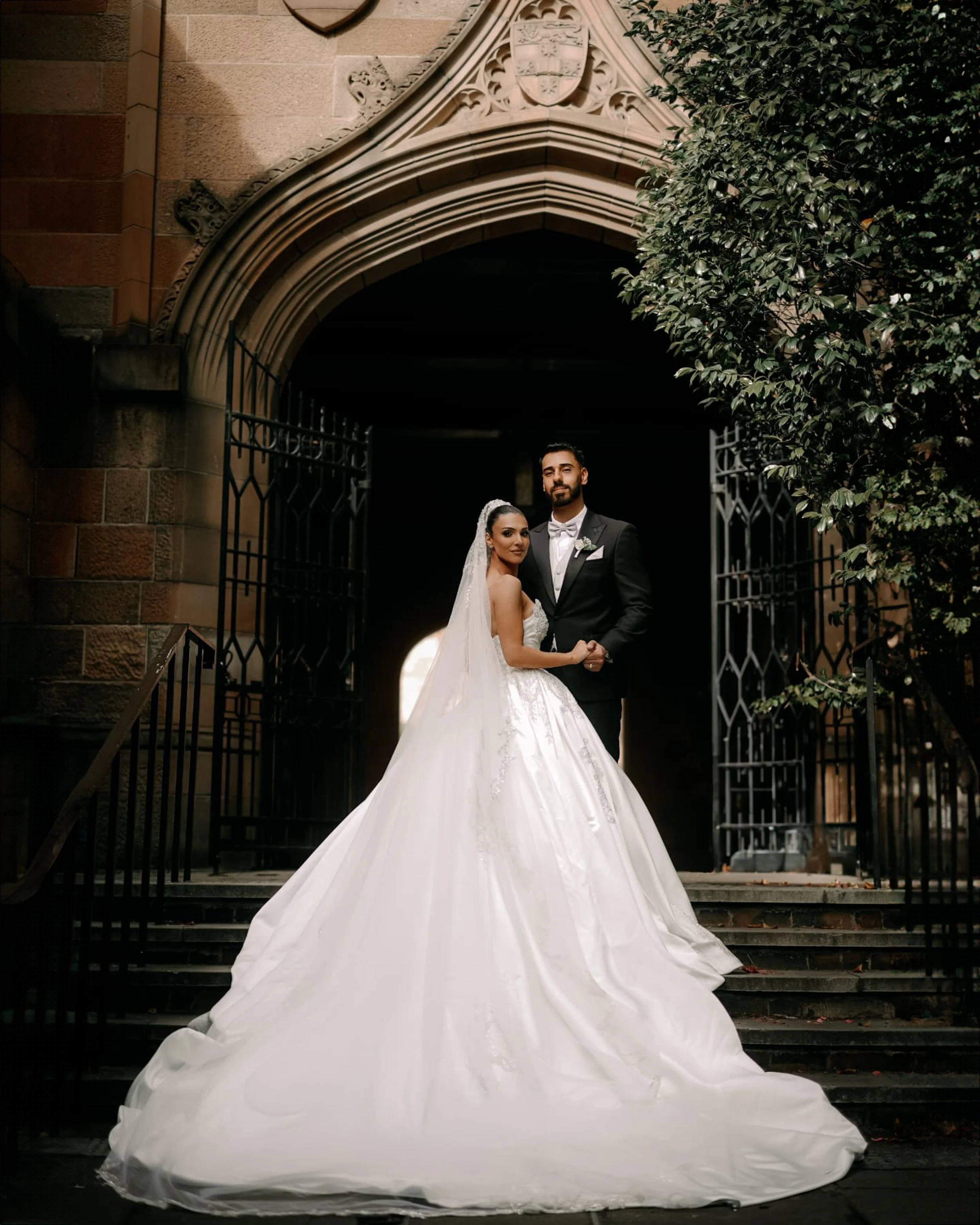 A bride and groom hold hands on steps in front of a Gothic-style archway, with the bride in a white wedding gown and veil, and the groom in a tuxedo, during their wedding photoshoot.