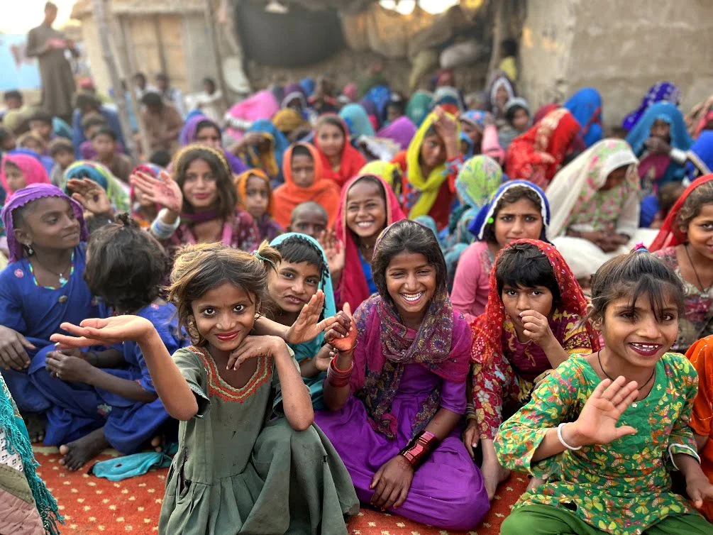 Children and women sitting outdoors on a red carpet, smiling and waving in colorful traditional clothing.